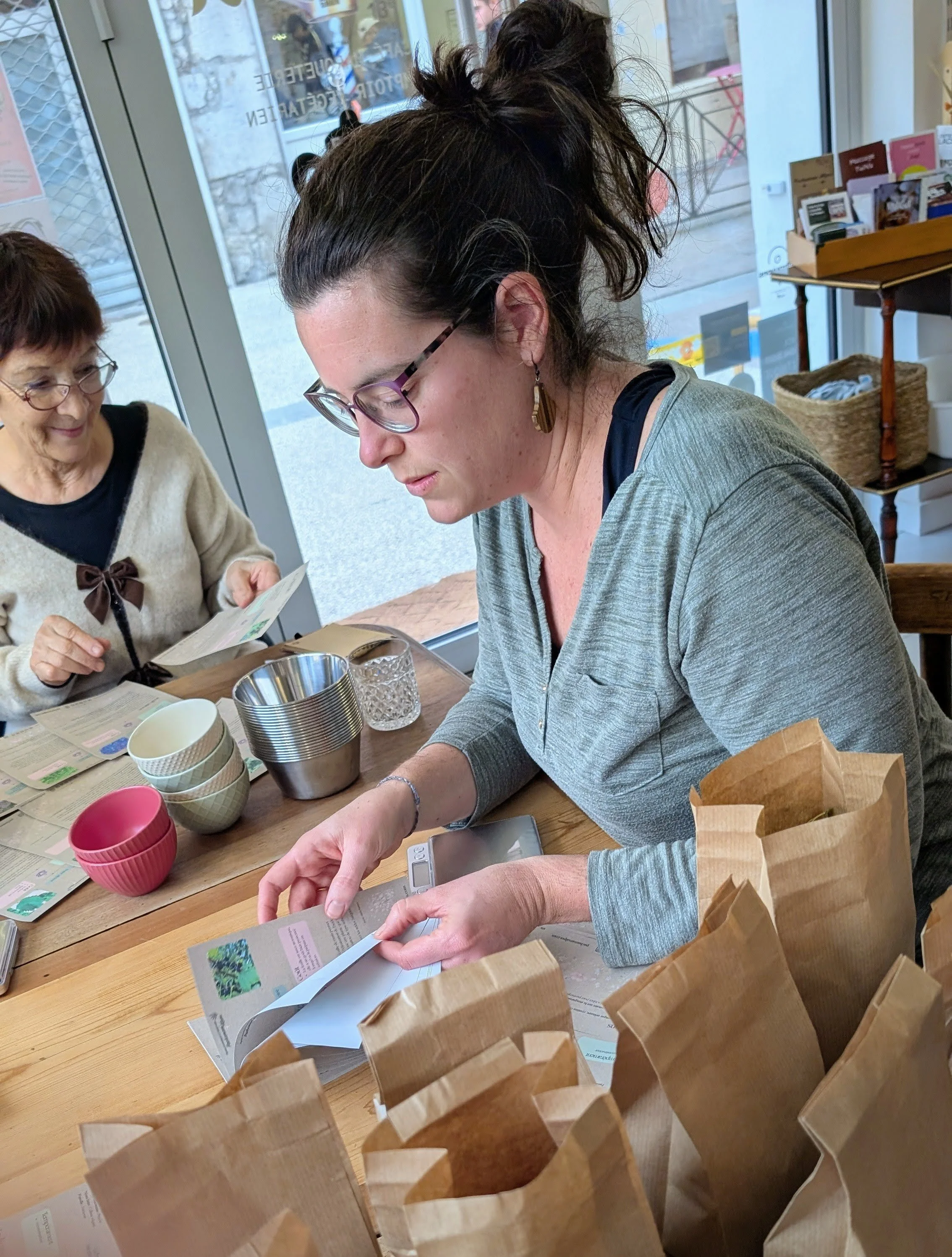 Une femme et une femme âgée assises à une table, la femme manipule une brochure ou un magazine, la femme âgée regarde un document. La table contient des tasses, un verre, des sacs en papier, et des objects divers. La scène se déroule près d'une grand