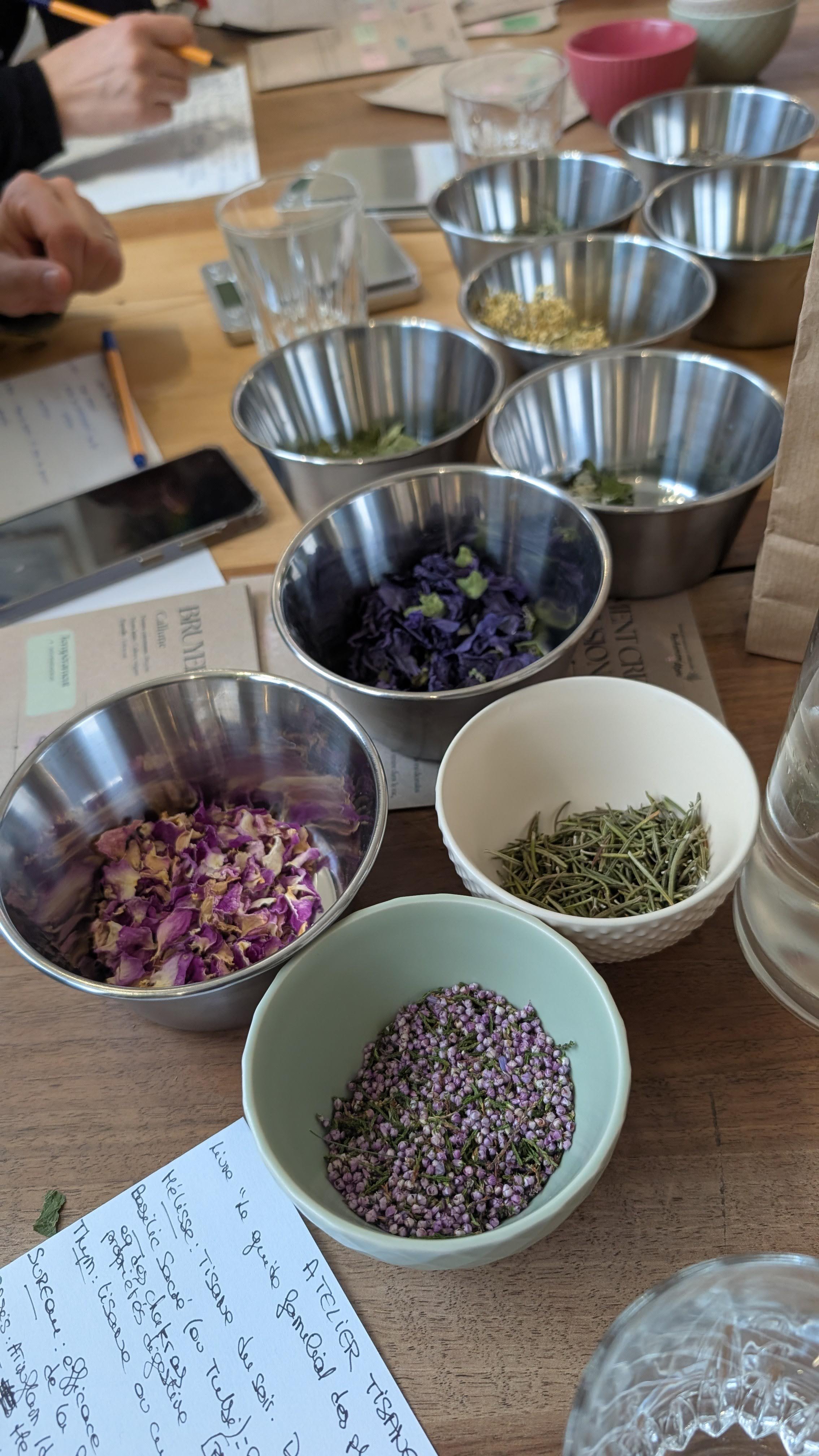 Différents types de fleurs séchées dans des bols en métal et en céramique, utilisées probablement pour des infusions ou des tisanes, sur une table avec papier et verre d'eau.