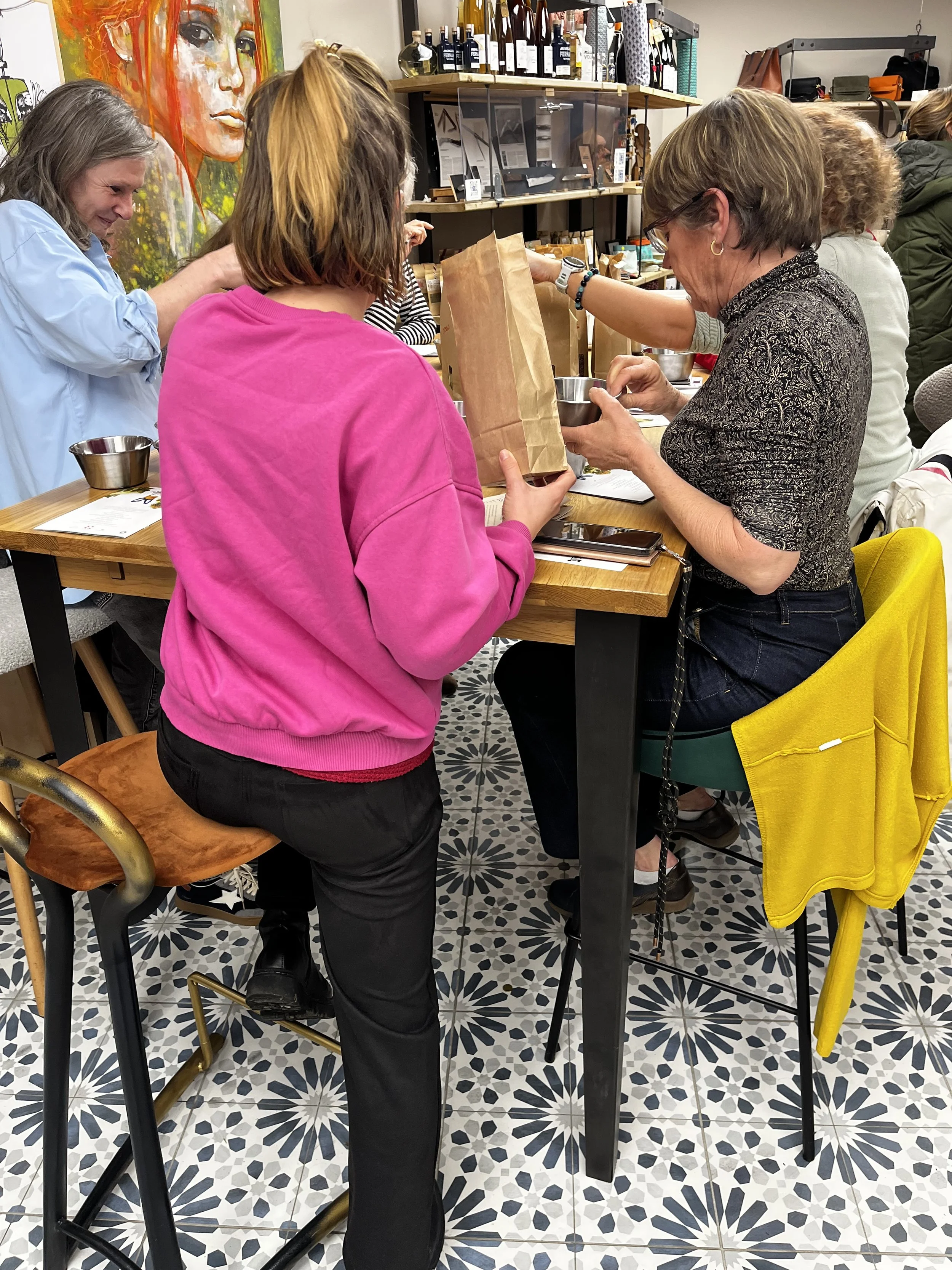 Groupe de personnes assises autour d'une table à un atelier ou une dégustation, échangeant avec des sacs en papier et des bols en acier.