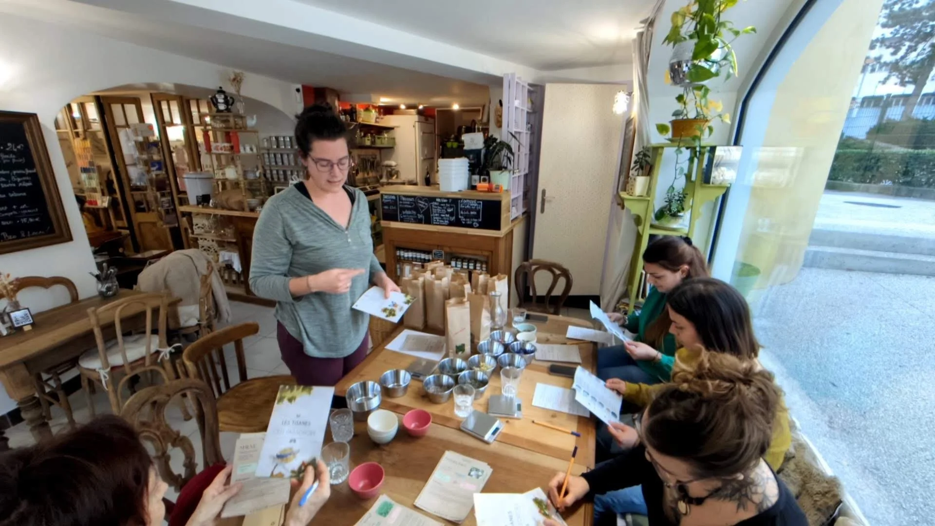 Femme expliquant un atelier à un groupe de personnes assises autour d'une table dans un café ou une boutique, avec divers contenants et documents.