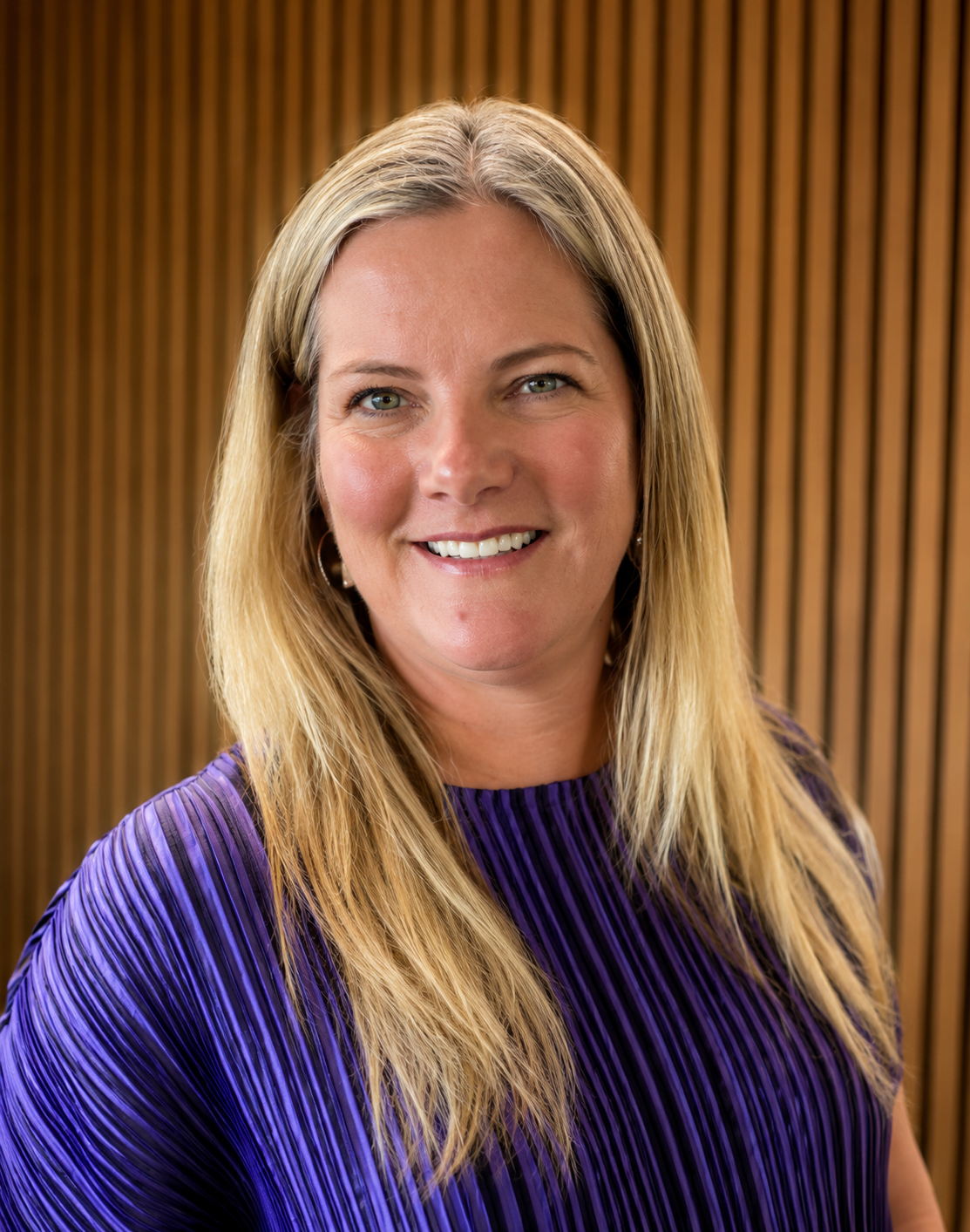 Close-up of a smiling woman with long blonde hair wearing a purple and black striped top, standing in front of a wooden slat background.