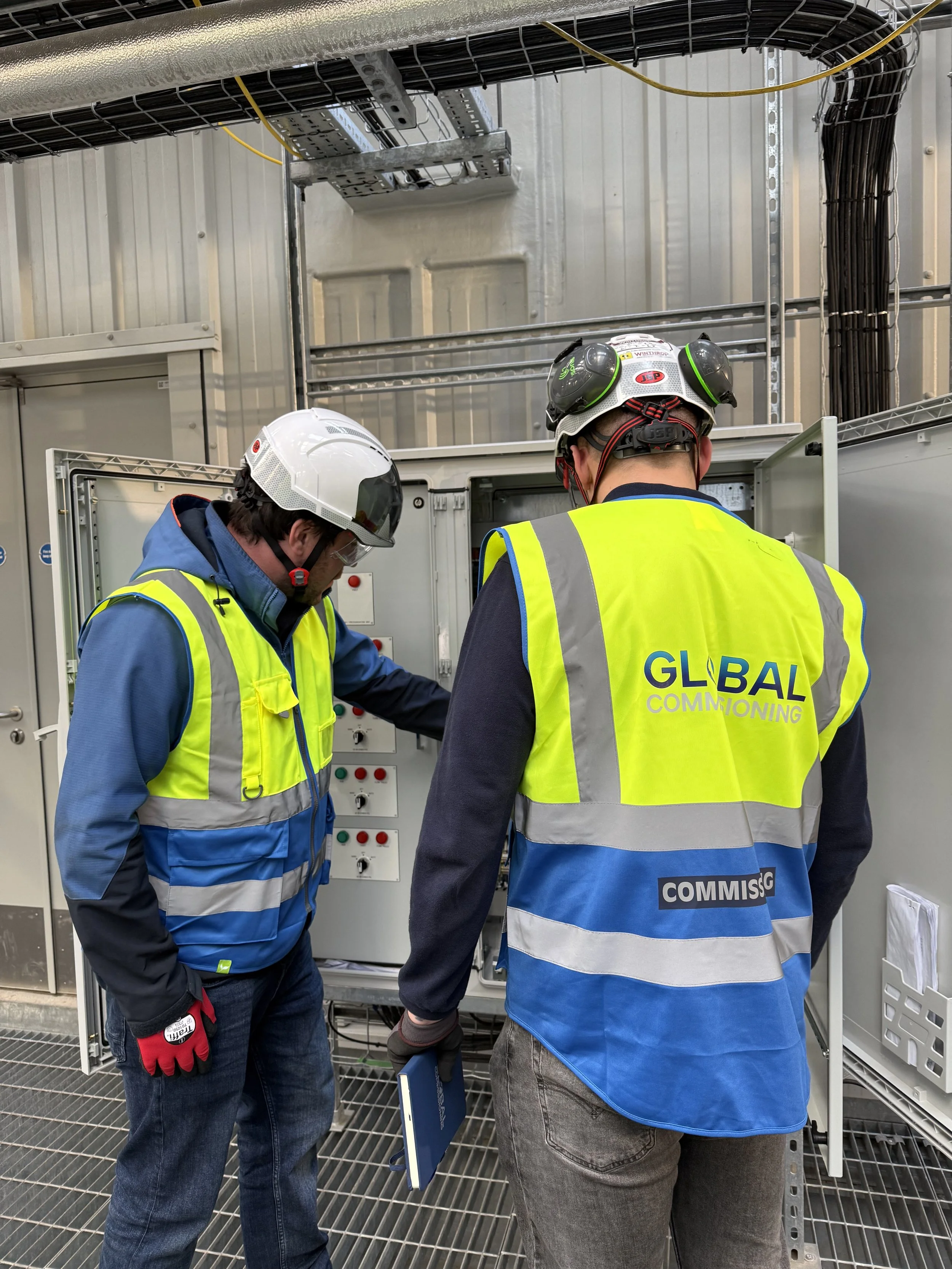 Two workers in safety gear inspecting electrical equipment inside a power distribution cabinet.