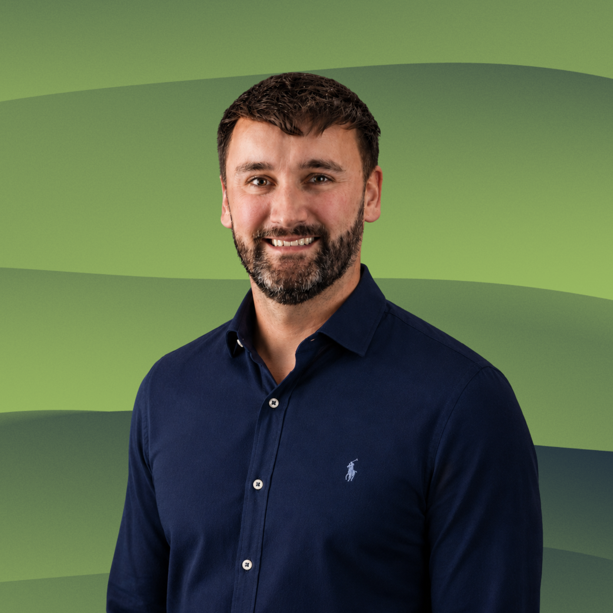 A smiling man with short brown hair, a beard, and blue eyes, wearing a navy blue button-up shirt with a small logo on the chest, standing in front of a wooden slat background.