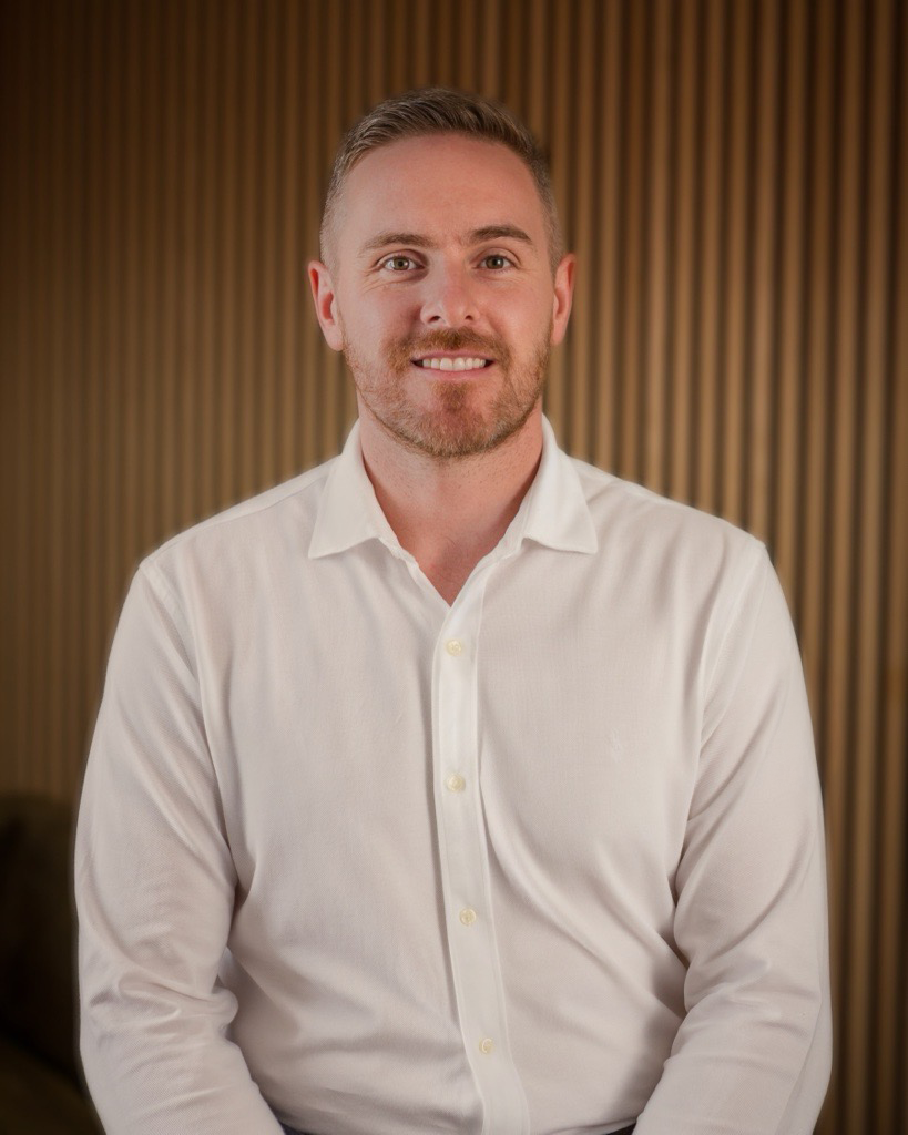 A man with short light brown hair and a beard, wearing a white dress shirt, smiling, standing in front of a wooden panel background.