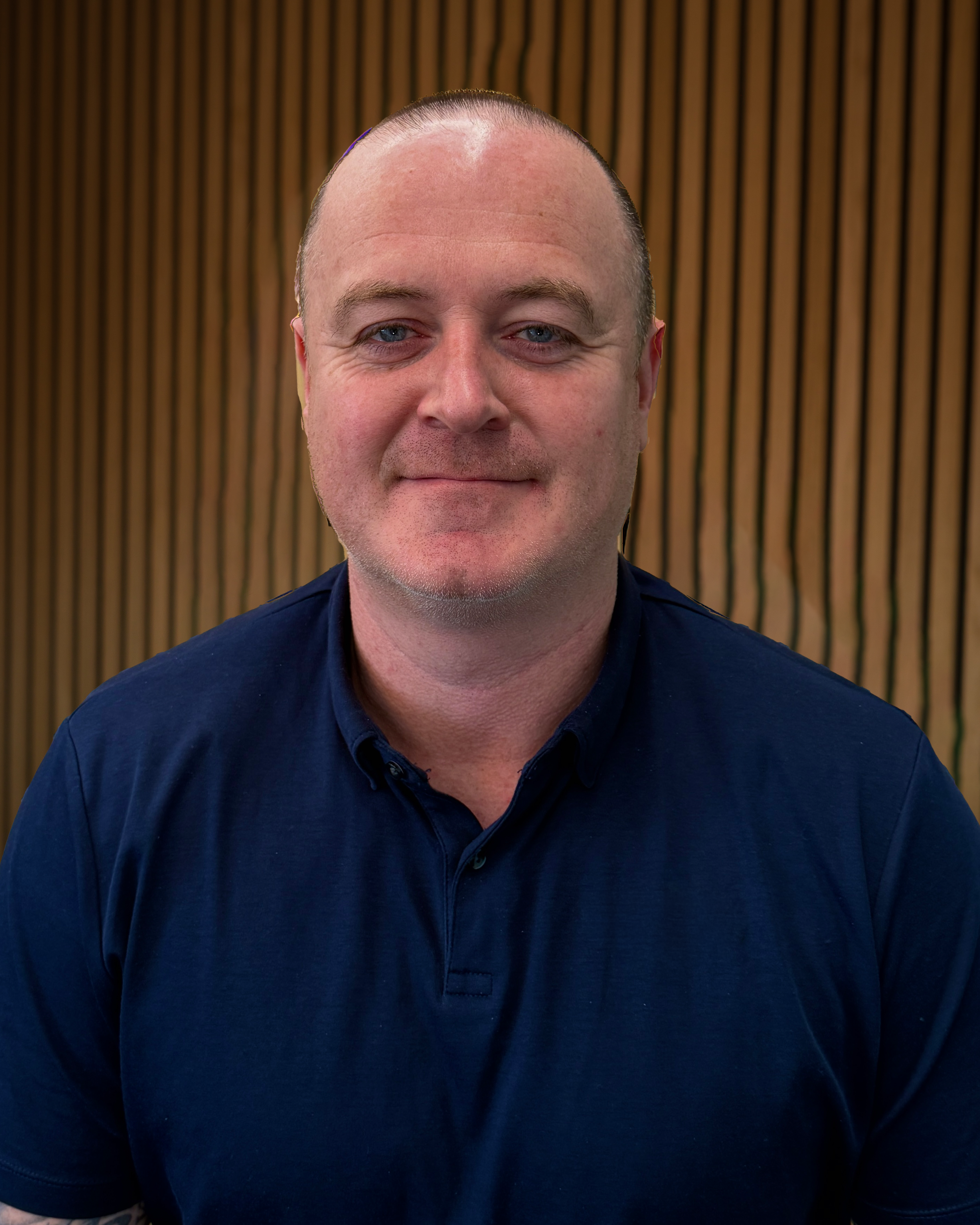 Close-up of a man with a shaved head, wearing a dark blue shirt, seated against a background of vertical wooden slats.