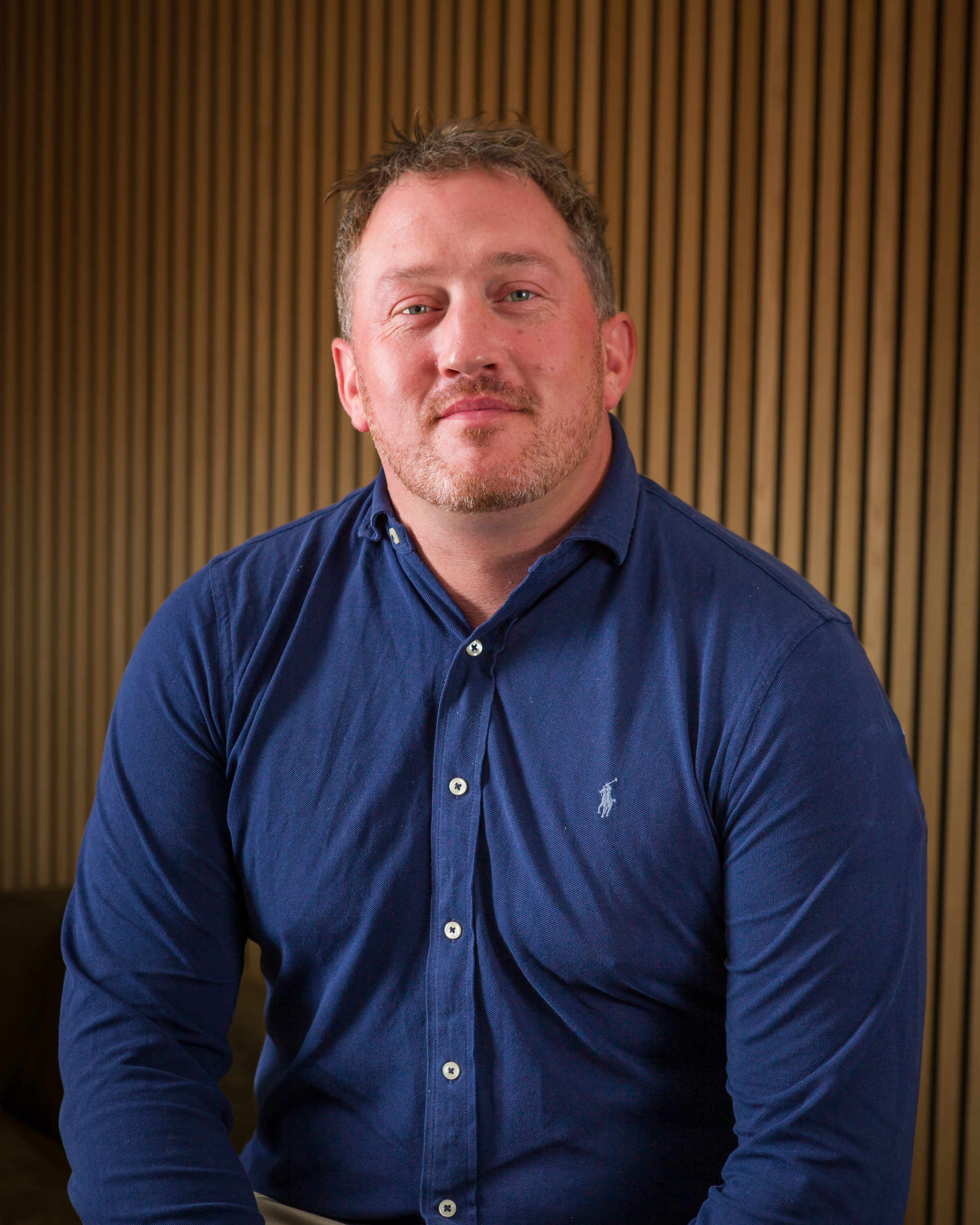 A man with light brown hair and a beard wearing a navy blue shirt, sitting against a wooden slat background.