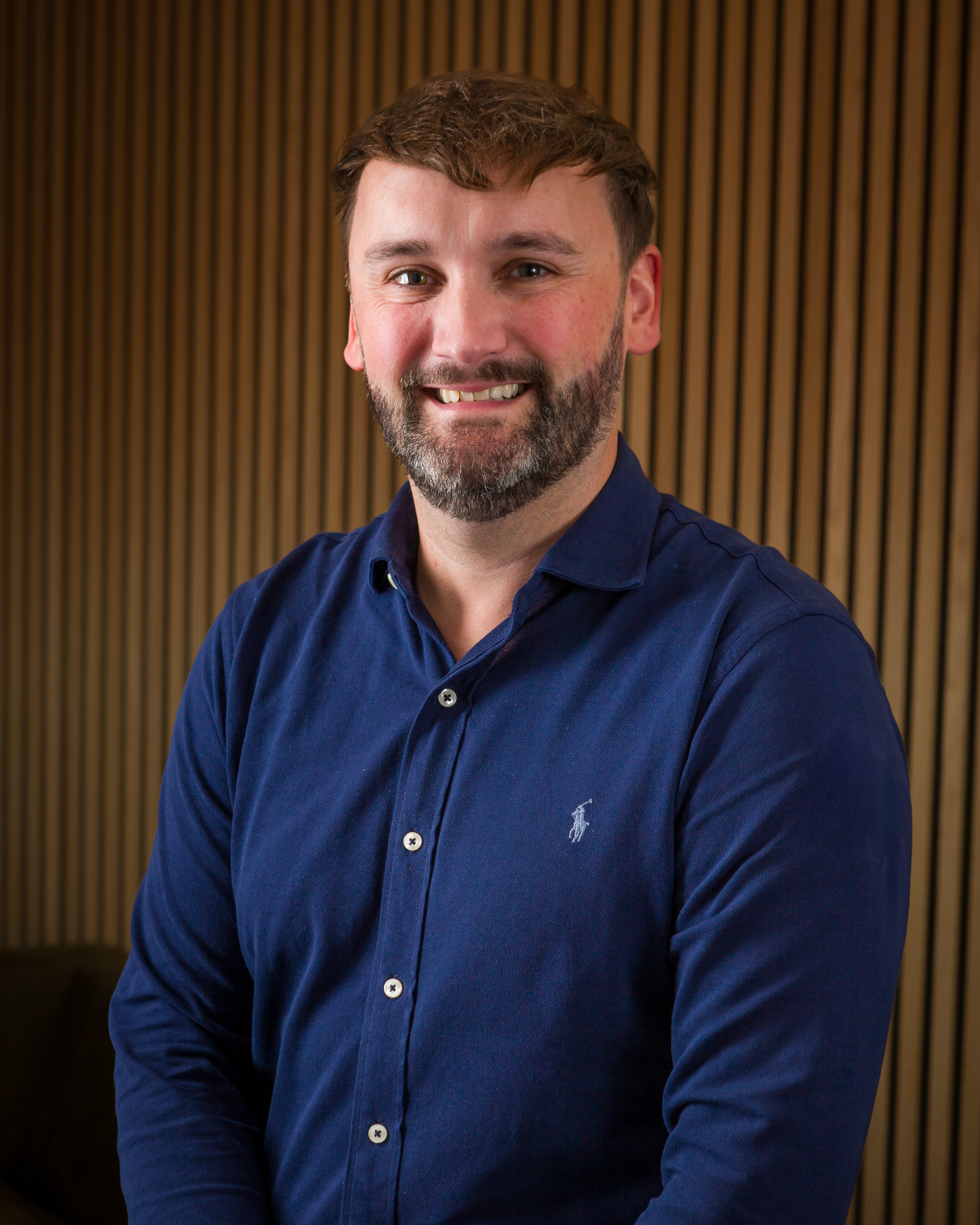 A smiling man with short brown hair, a beard, and blue eyes, wearing a navy blue button-up shirt with a small logo on the chest, standing in front of a wooden slat background.