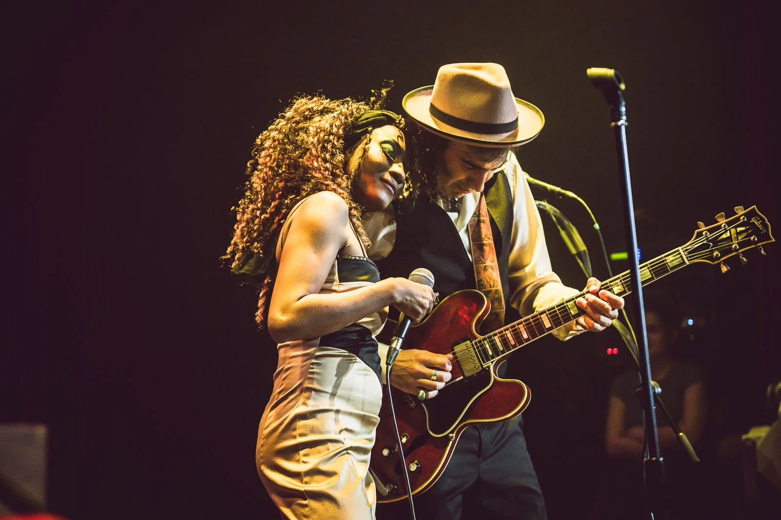 Una mujer y un hombre tocando música en un escenario, con la mujer sosteniendo un micrófono y el hombre tocando una guitarra eléctrica.