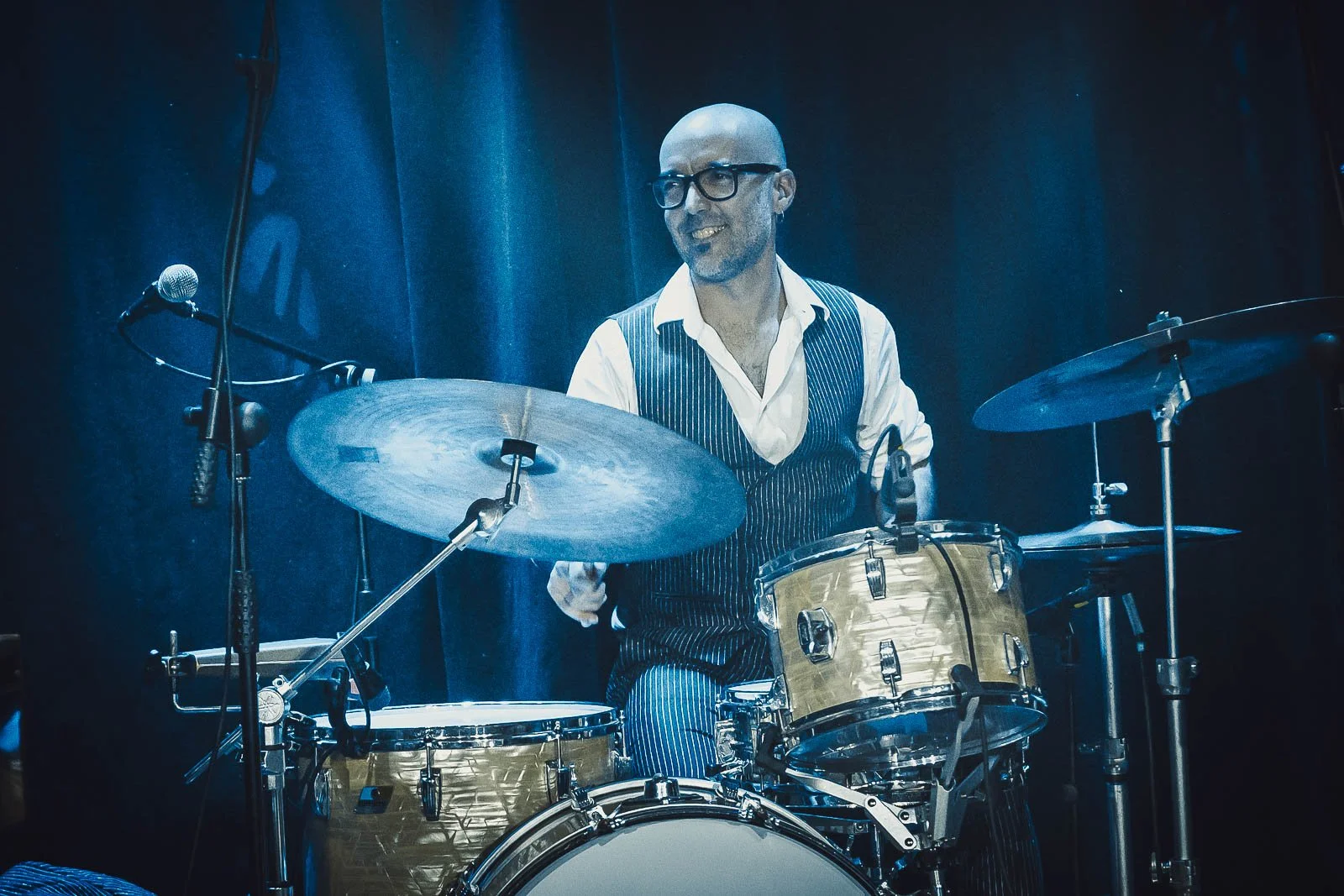 Hombre tocando la batería en un escenario, vestido con chaleco a rayas y gafas, con cortos en la cabeza y barba, fondo oscuro
