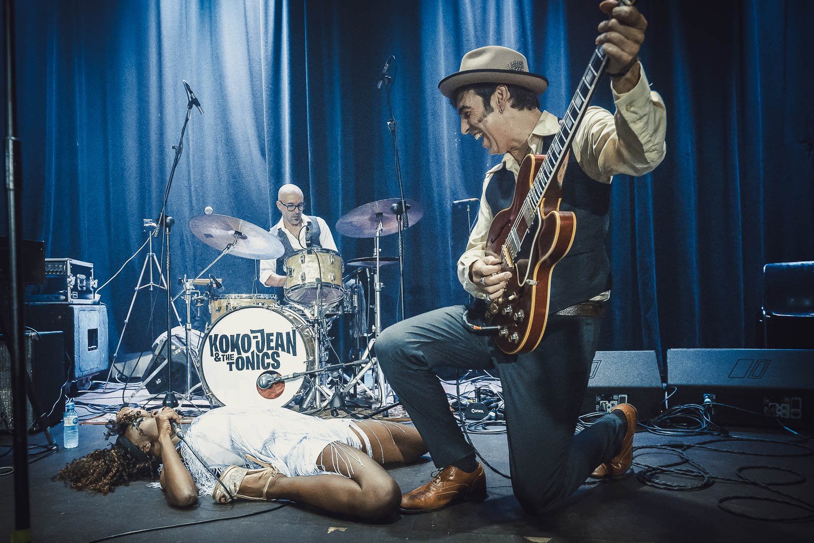 Una banda tocando música en un escenario con una mujer en el suelo, un hombre con guitarra y otro con batería.