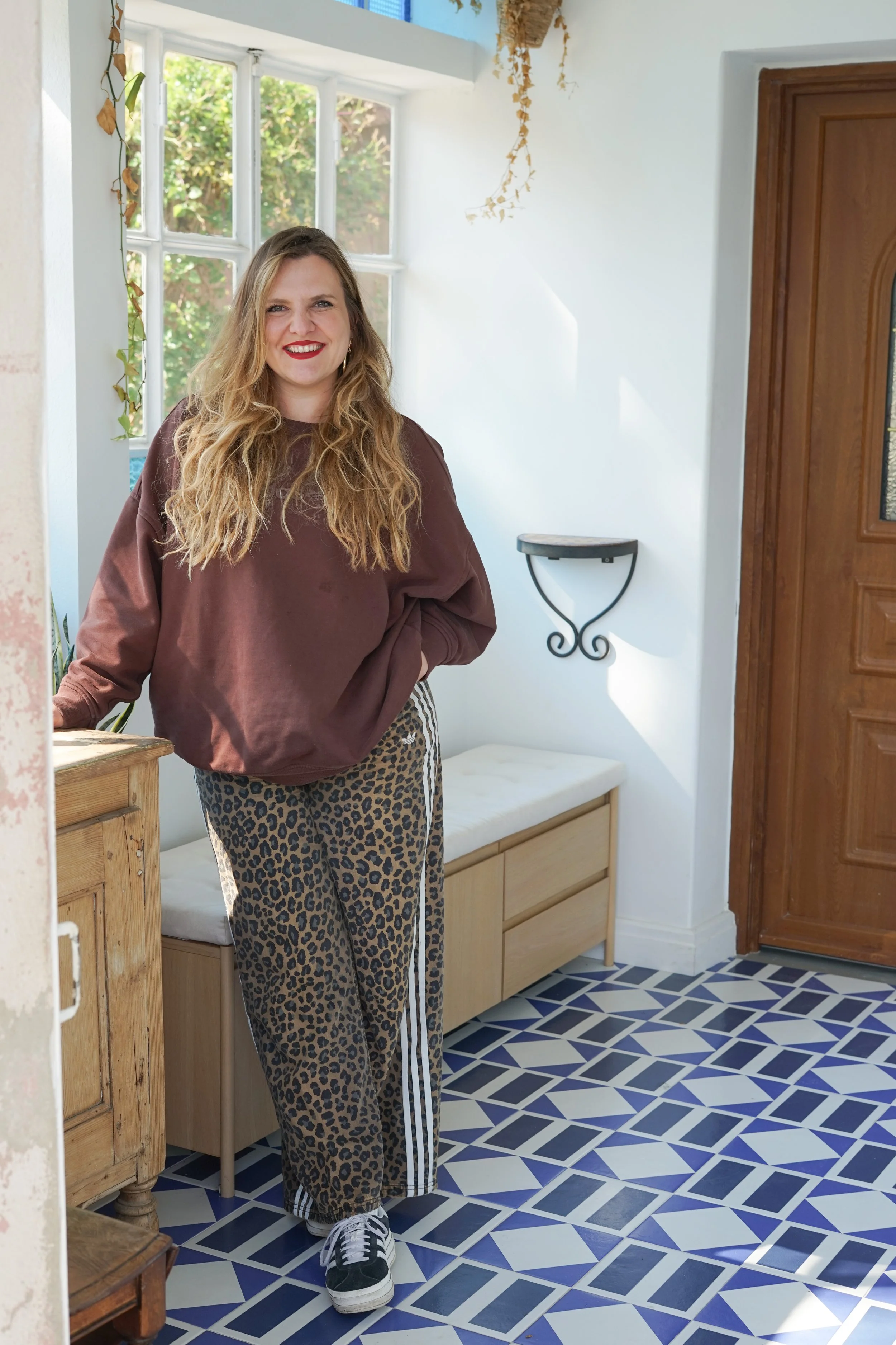 Femme avec long cheveux blonds, portant un sweat marron, un pantalon à motif léopard et des baskets noires et blanches, souriante, à l'intérieur d'une pièce lumineuse avec un sol en carreaux bleus et blancs, et un grand volet en bois.