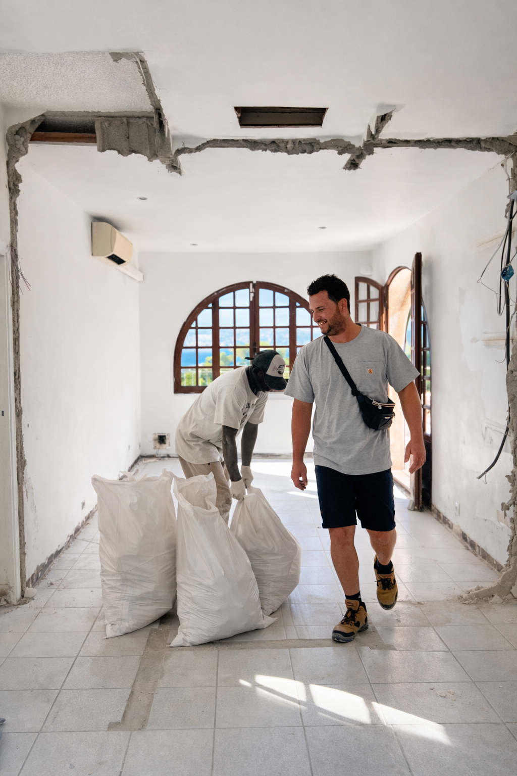 Deux hommes rénovant une pièce, un souriant avec un sac à bandoulière, l'autre portant des sacs de matériaux, dans une pièce lumineuse avec une grande fenêtre en arc.