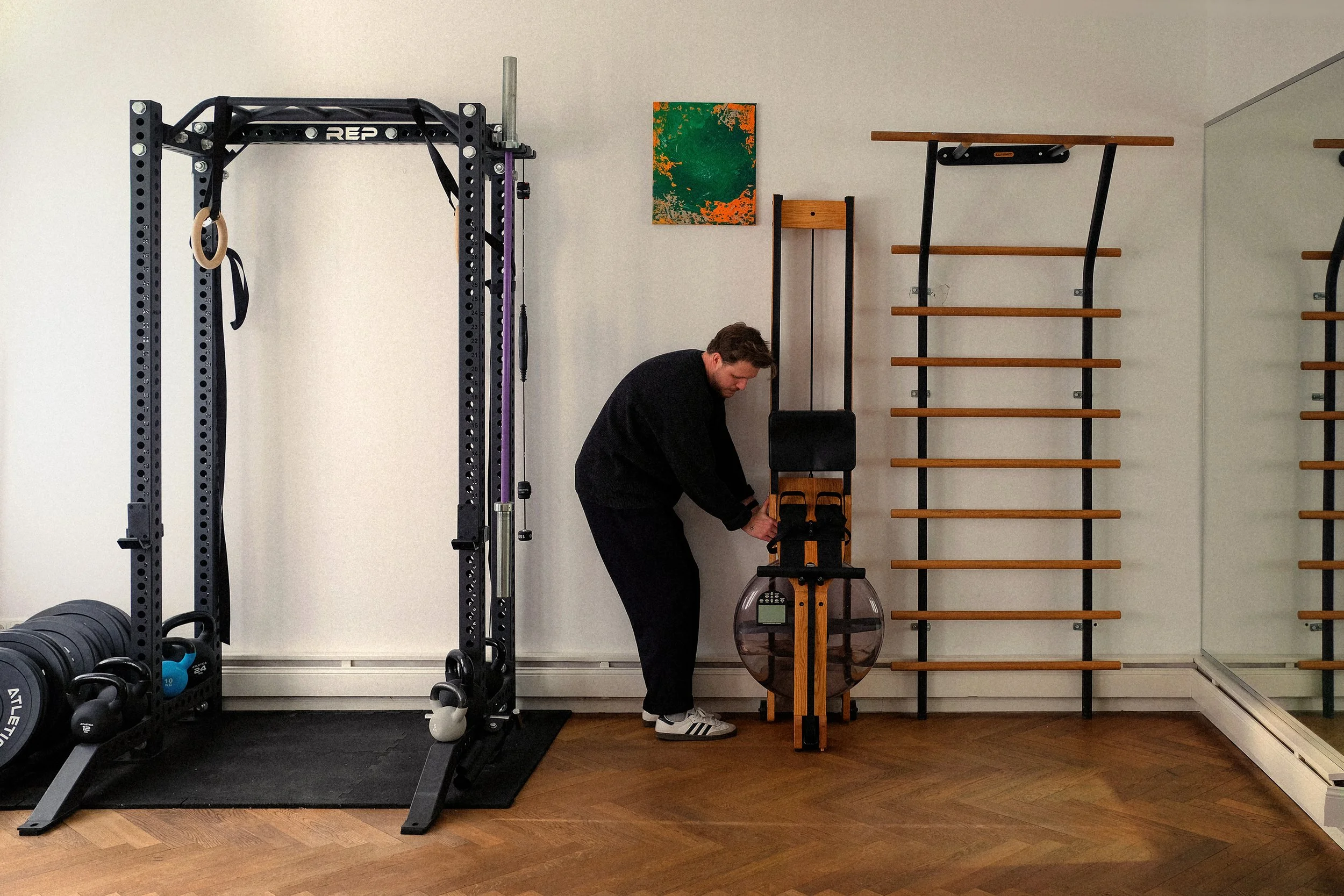 Physiotherapeut Darius Münch steht im hinteren Raum der Praxis. Links neben ihm sind ein Krafttrainingsrack und Kettlebells, rechts ein Rudergerät und ganz rechts eine Klimmzugstange.