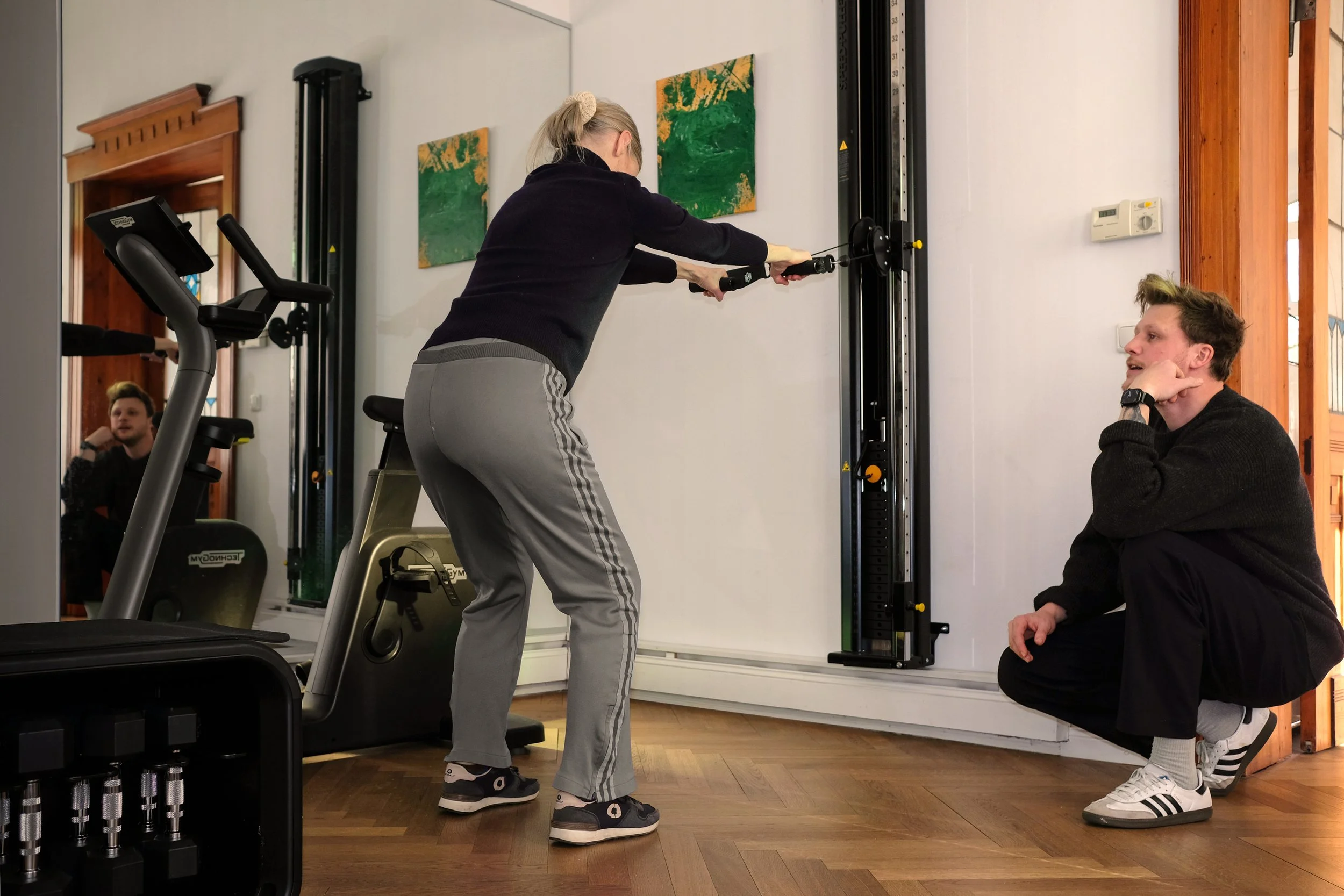 Frau in den 50ern trainiert an einem Kabelzug, während Darius Münch, der Physiotherapeut, sie daneben berät.