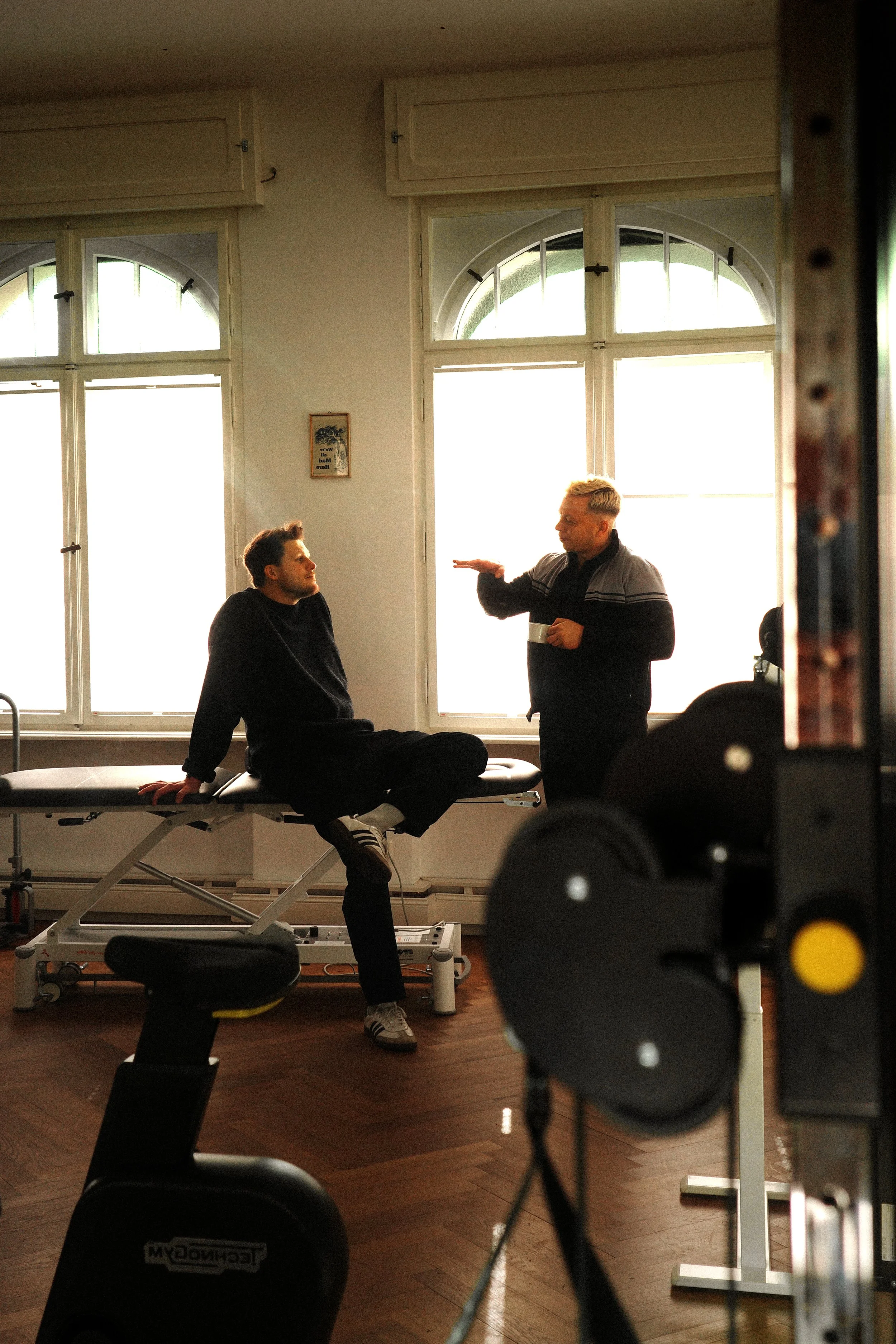 Zwei Physiotherapeut*innen unterhalten sich locker in einer Praxis, einer steht mit einer Tasse in der Hand, der andere sitzt auf einer Behandlungsliege, weitere Fitnessgeräte im Vordergrund.