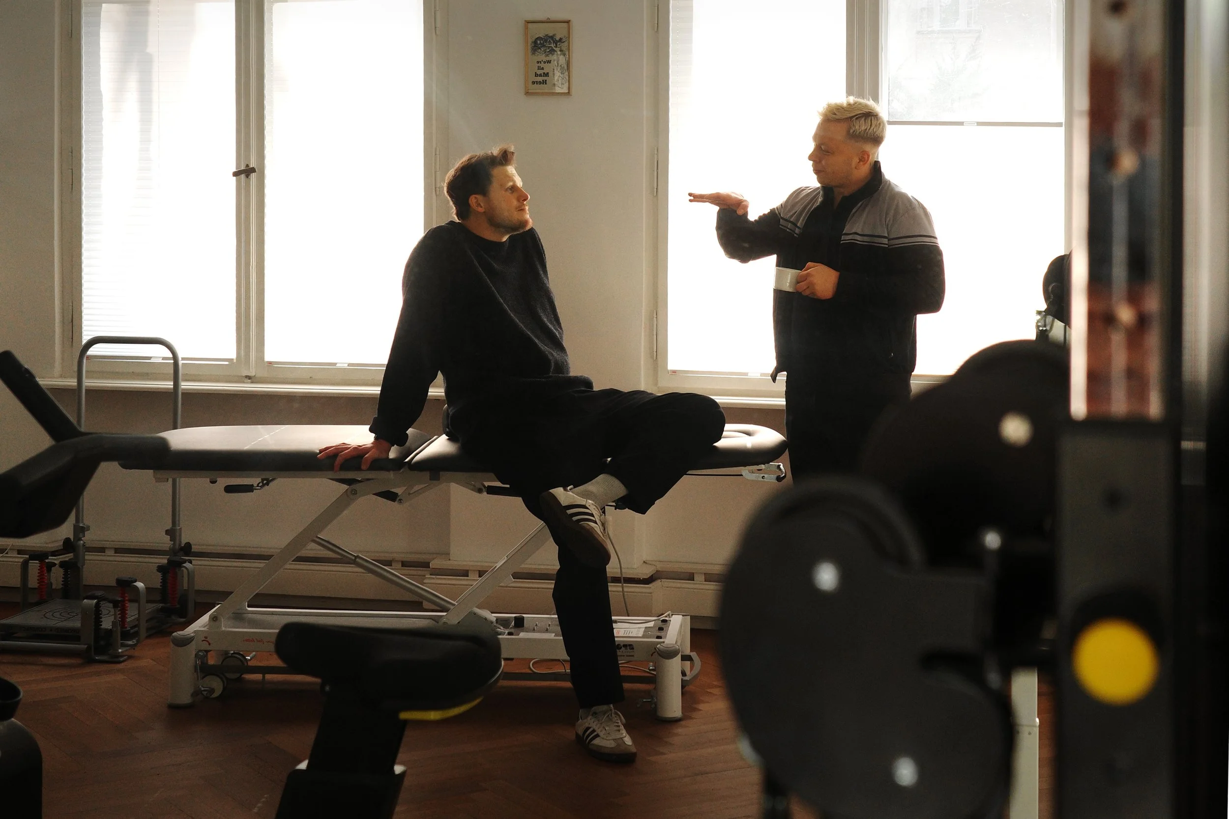 Weiterer Blick in den hinteren Raum der Physiotherapiepraxis. Darius Münch sitzt auf der Behandlungsliege und spricht mit einem Kollegen, im Hintergrund steht ein sensomotorisches Therapiegerät.