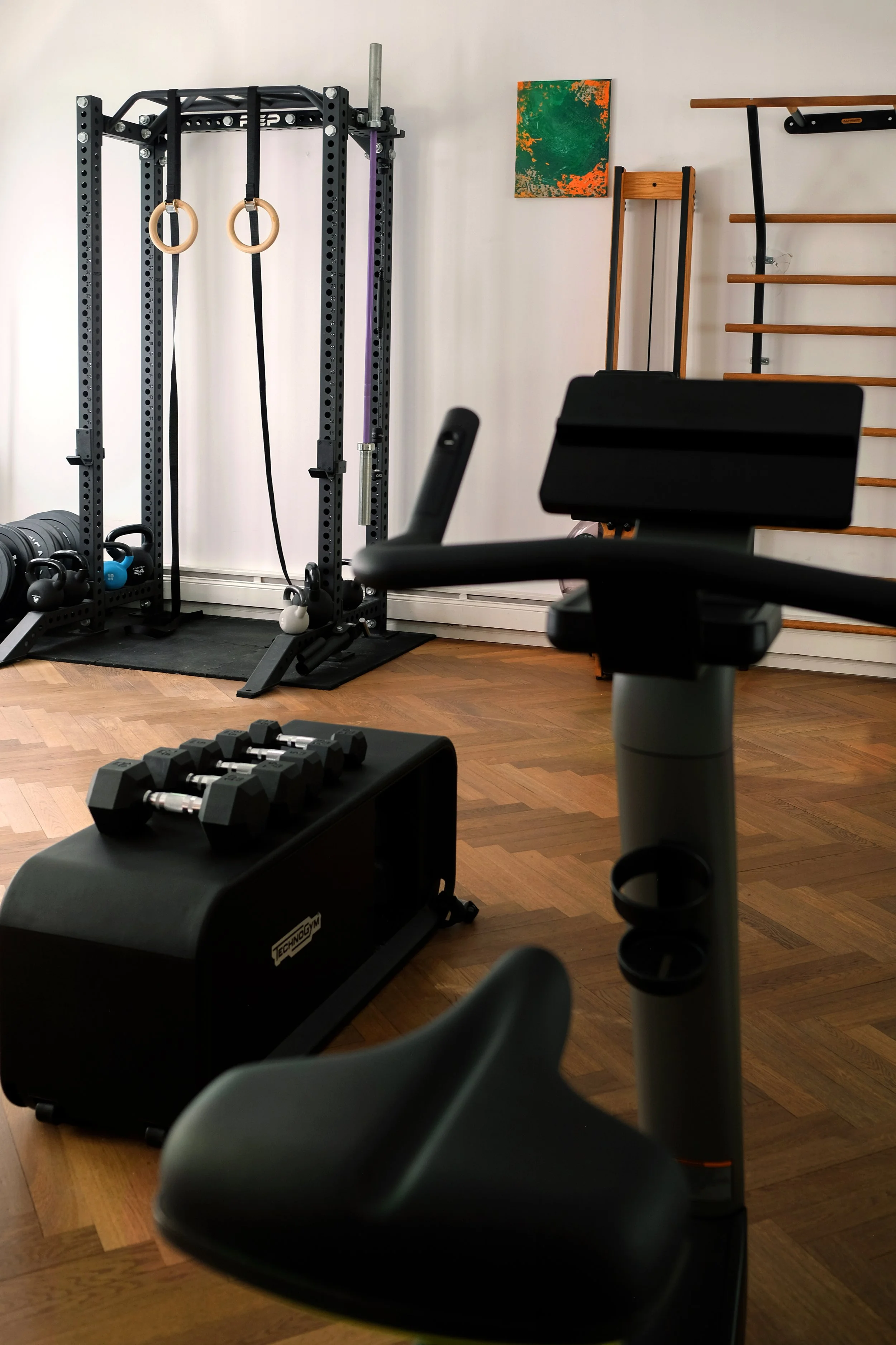 Weiterer Blick in den hinteren Raum der Physiotherapiepraxis. Im Vordergrund steht ein Ergometer, im Fokus sind Hanteln und eine Bank, im Hintergrund ein Krafttrainingsrack, ein Rudergerät und eine Klimmzugstange.