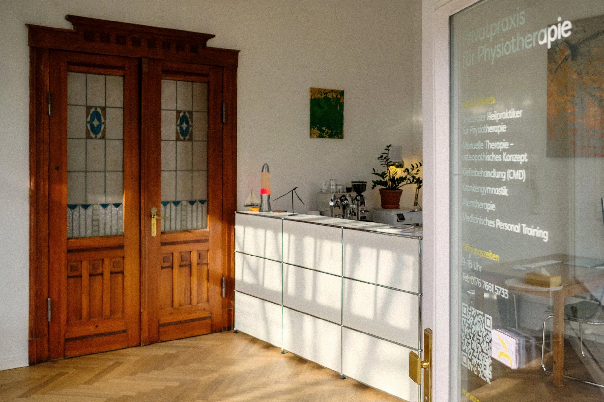 Vorderer Raum der Physiotherapiepraxis mit Kaffeestation und Beratungsbereich im hinteren Teil. In der Mitte steht eine Reihe weißer Schränke, rechts führt eine alte Holztür mit Buntglas in den hinteren Praxisraum.