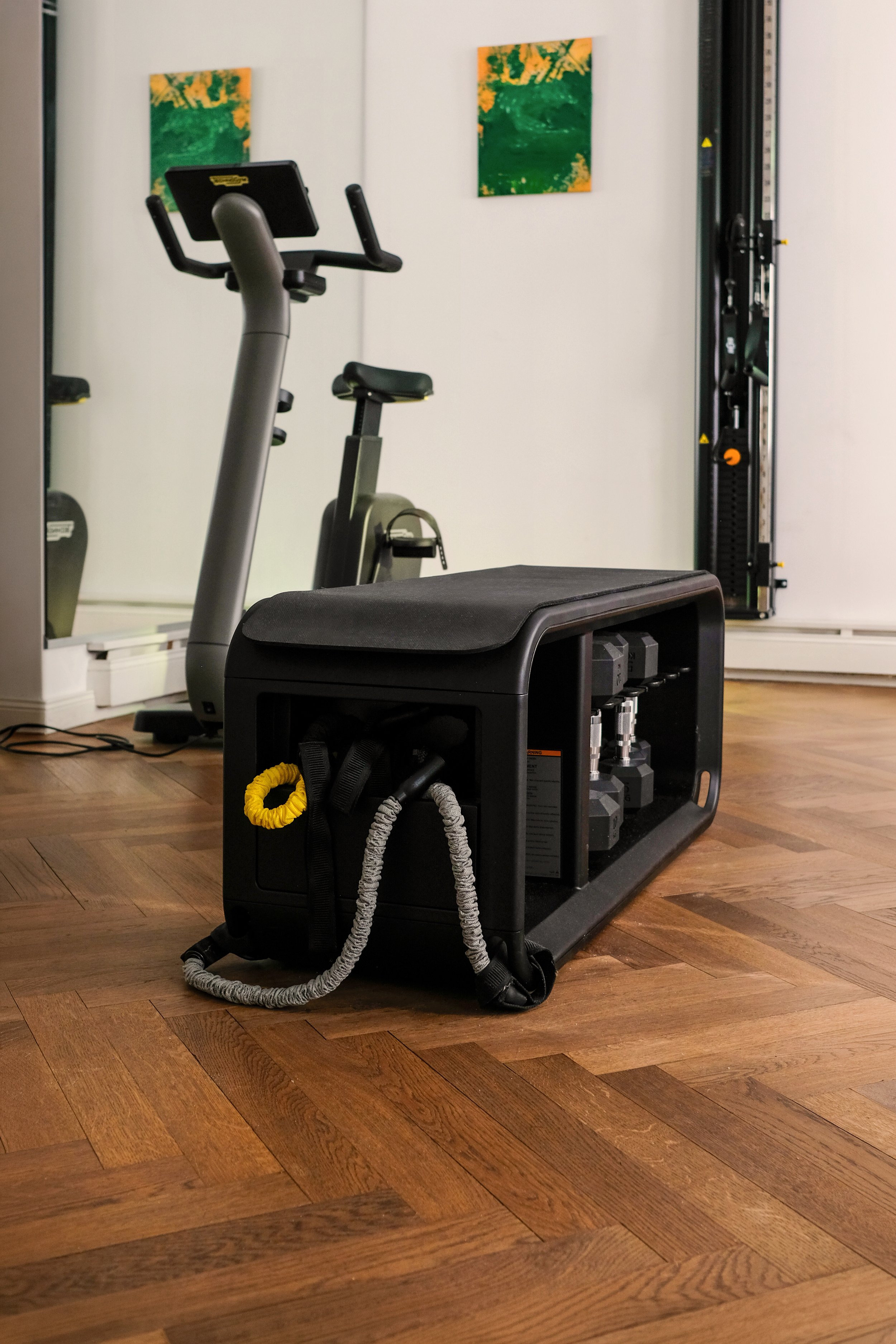 Weiterer Blick in den hinteren Raum der Physiotherapiepraxis. Im Fokus sind Hanteln und eine Bank, im Hintergrund ein Ergometer, ein Kabelzug und ein großer Spiegel.