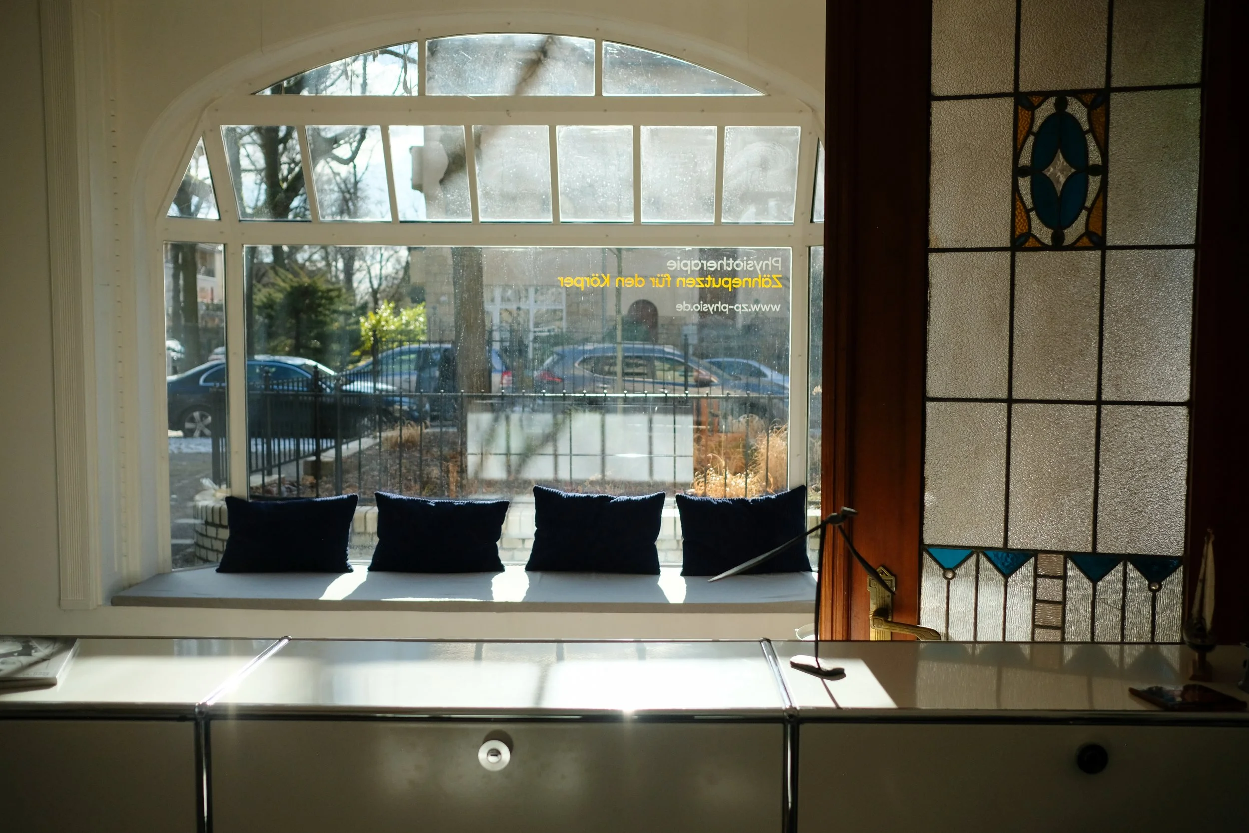 Blick durch das große Fenster im vorderen Raum der Physiotherapiepraxis auf die Straße. Die Sonne scheint auf die Kissen der Sitzbank auf der Fensterbank.