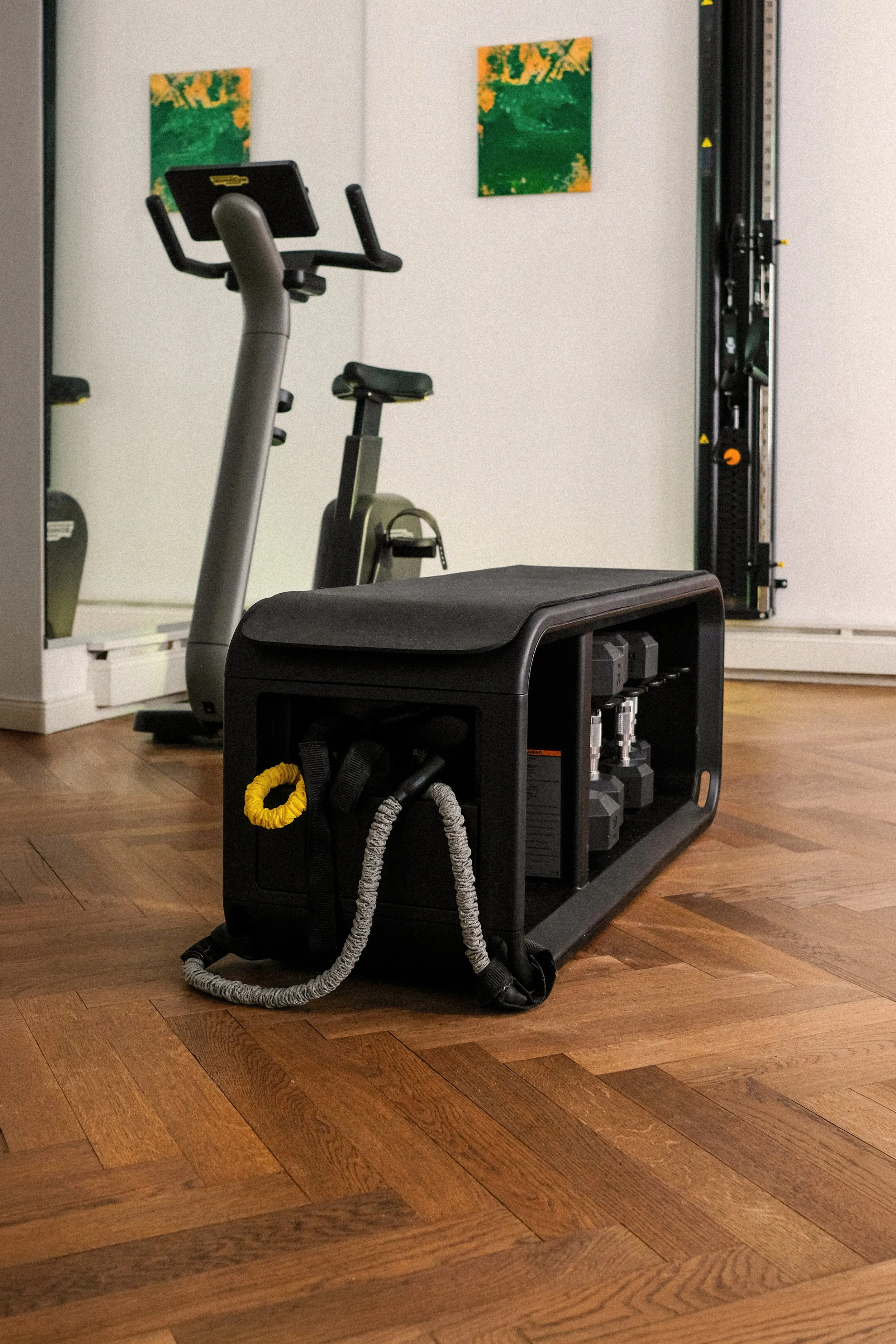 Weiterer Blick in den hinteren Raum der Physiotherapiepraxis. Im Fokus sind Hanteln und eine Bank, im Hintergrund ein Ergometer, ein Kabelzug und ein großer Spiegel.