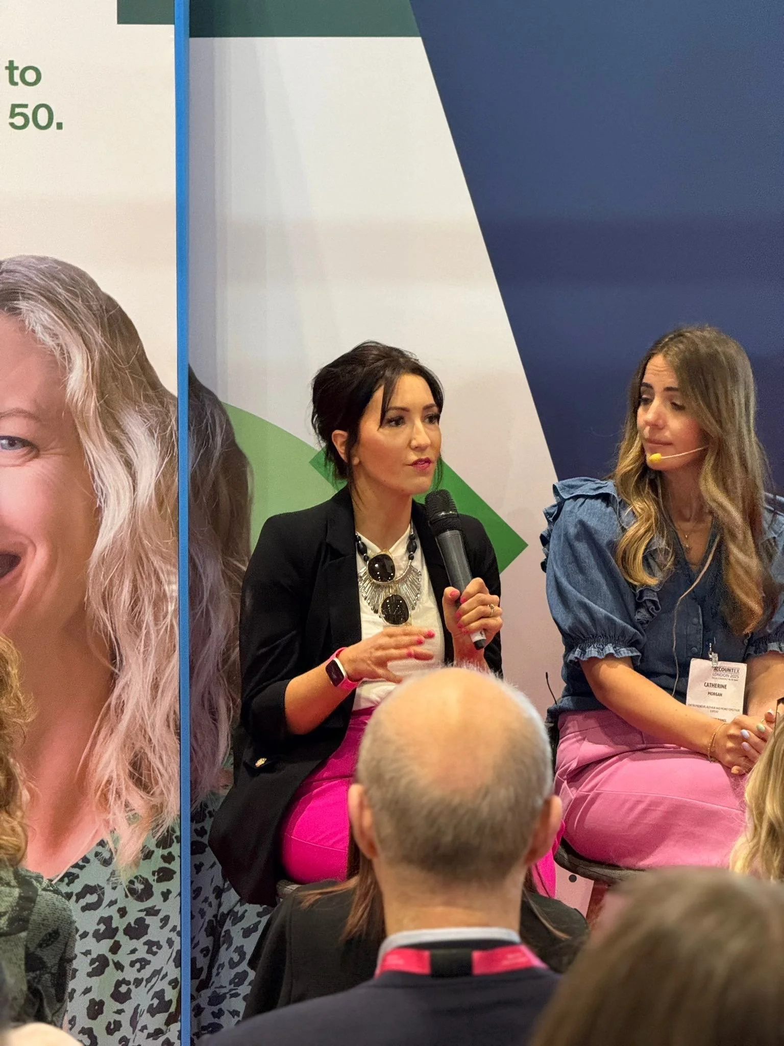 Two women sitting on a stage, one holding a microphone and speaking, during a panel discussion at a conference or event. The woman on the right is wearing a denim jacket and pink pants, while the woman on the left is wearing a black blazer with large sunglasses hanging from her neck.