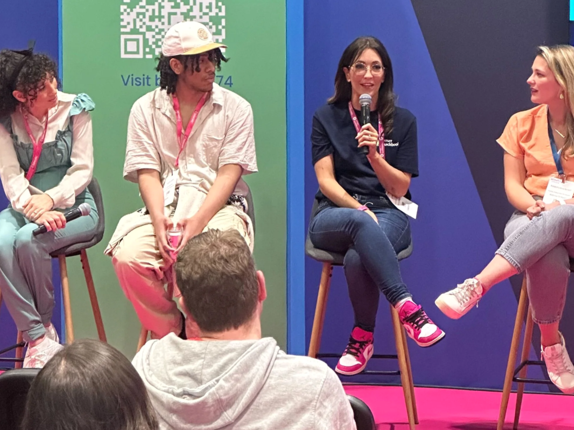 Four people are sitting on stools on stage at a panel or discussion event. A woman with dark hair and glasses is holding a microphone. To her right, a woman in a light orange shirt is speaking. To her left, a man with dreadlocks and a cap, and a woman with curly hair are listening. The background features a blue and green backdrop with a QR code and some text.