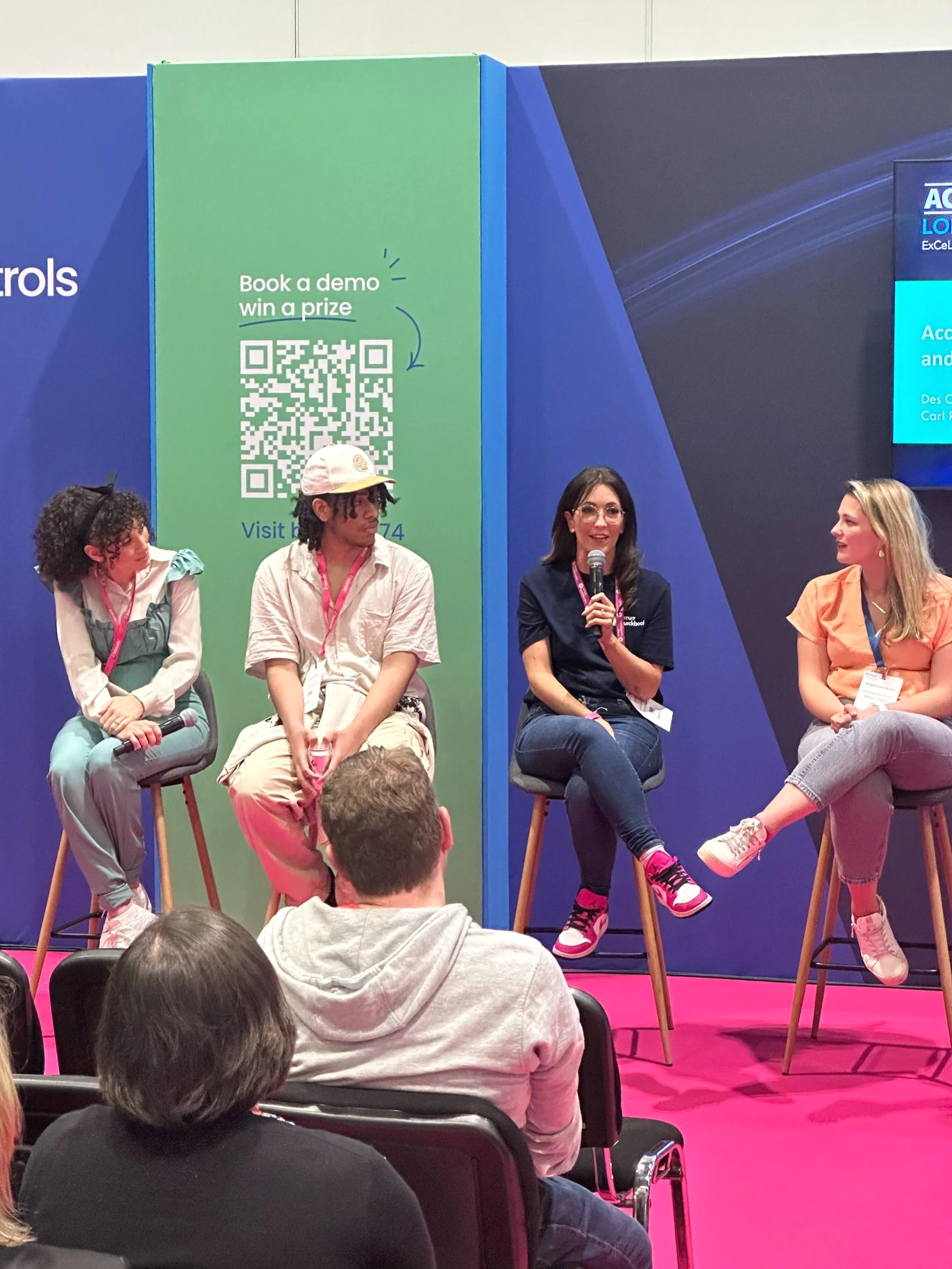 Four people sit on stools on a stage, participating in a panel discussion at a conference. A woman with glasses and a dark shirt holds a microphone while talking. The other three are listening, with one man wearing a cap, a woman with curly hair, and a woman in a peach shirt. Behind them is a large banner advertising a demo contest with a QR code. Audience members are seated in front of the stage.