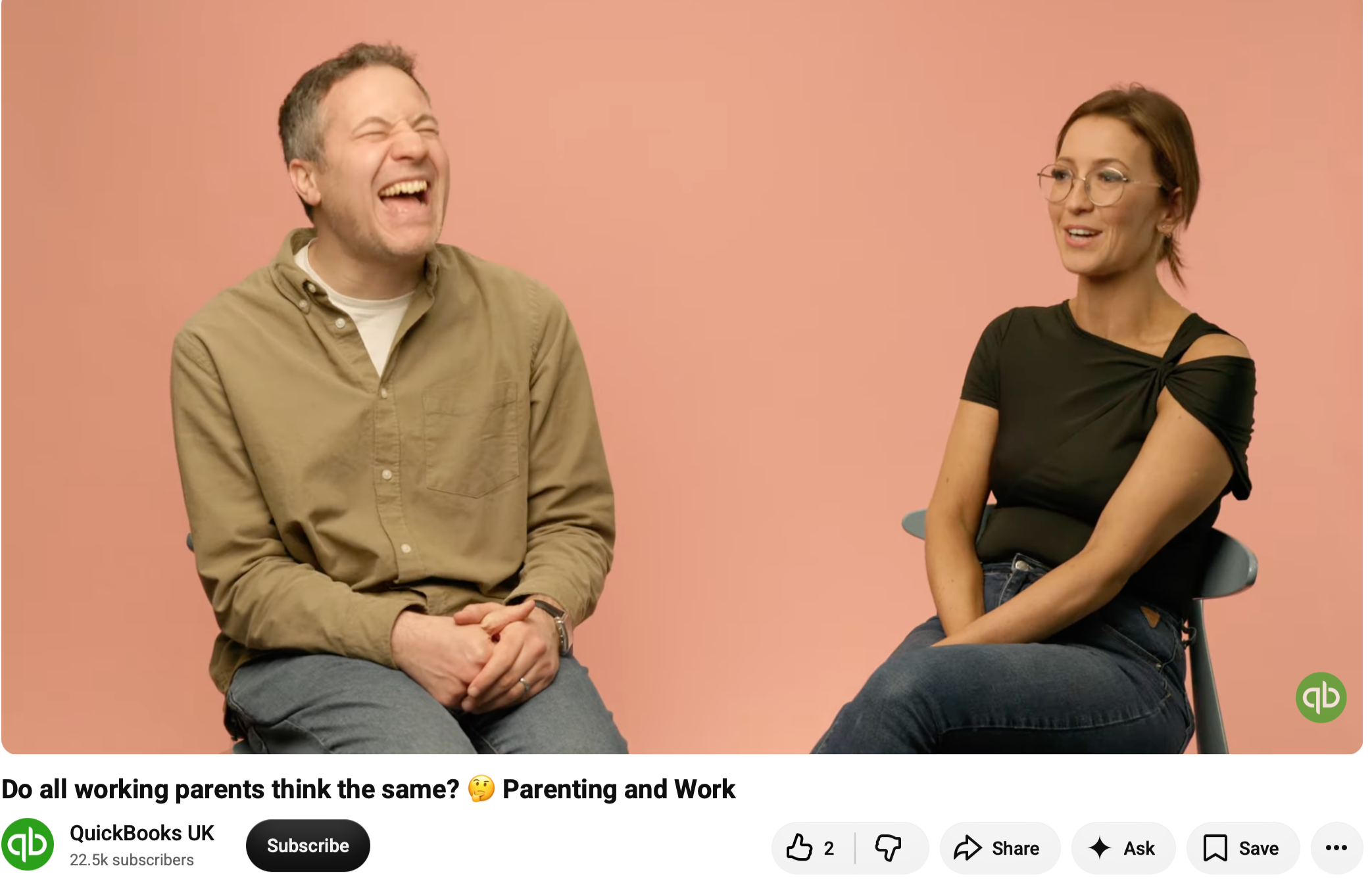 A man and a woman sitting on chairs against a pink background, laughing and having a conversation.