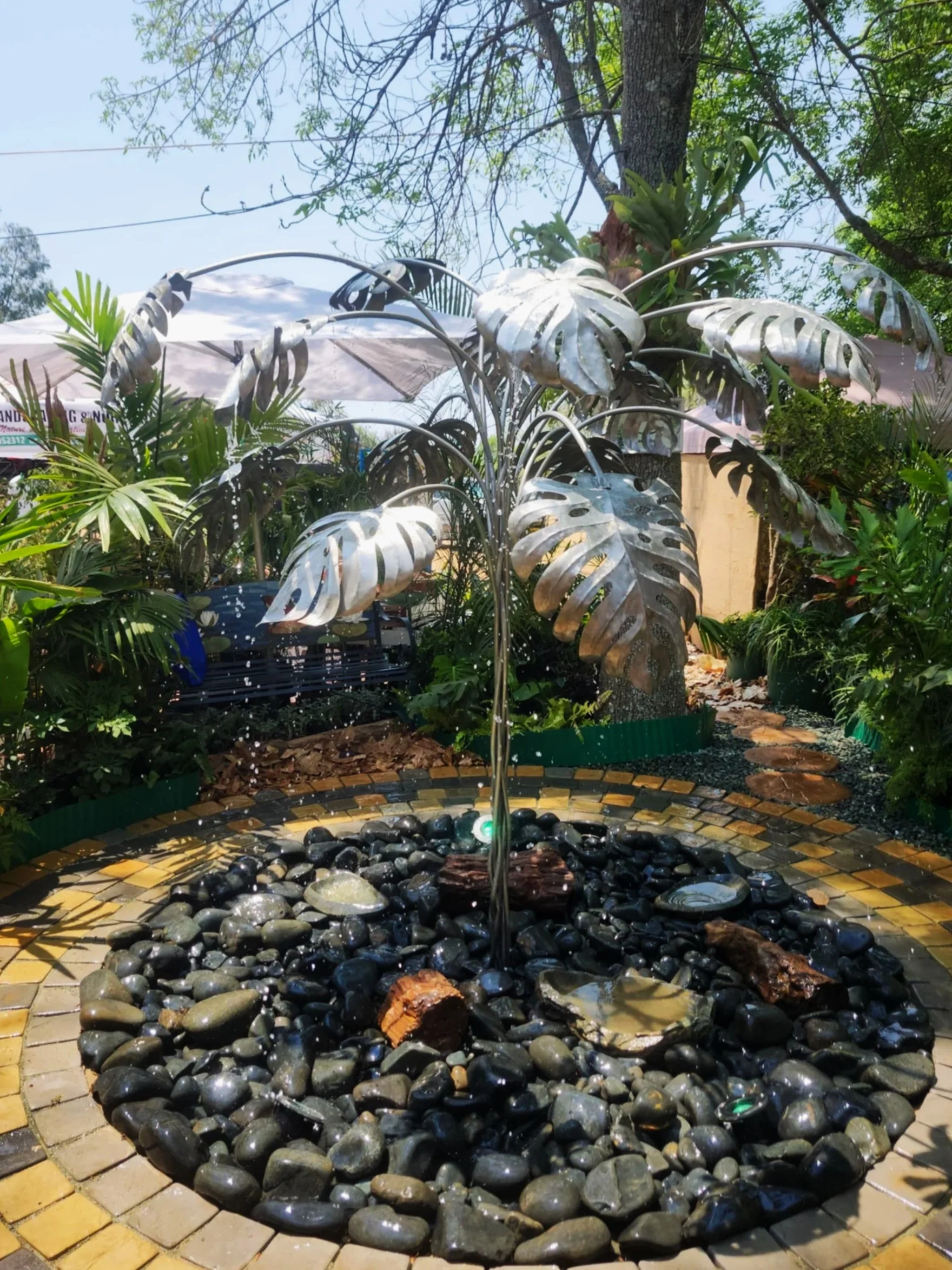 A decorative garden water fountain with a metallic Monstera leaf sculpture on top, surrounded by black and gray stones, with green plants and trees in the background.