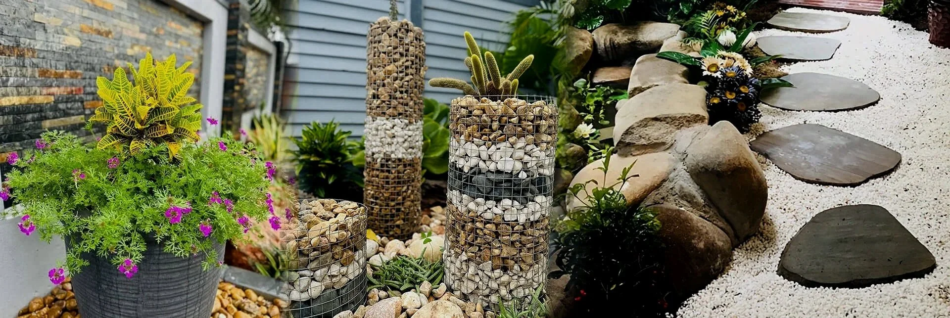 A garden with potted plants, tall cylindrical wire cages filled with stones and cacti, a stone pathway with large flat stones, and a rock border with flowers and greenery.