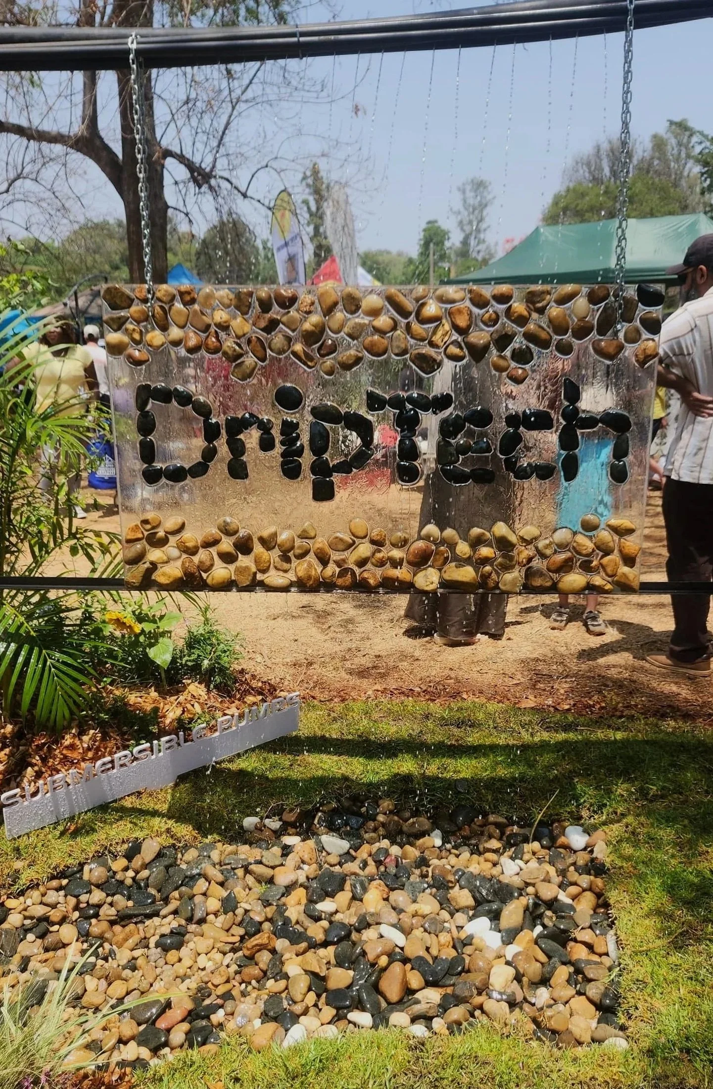 Sign with the word 'DRIPTECH' written using black pebbles, surrounded by a border of small mixed pebbles, at an outdoor event with people and tents in the background.