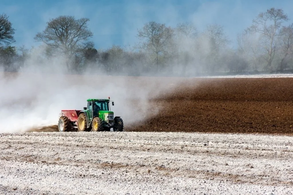 A green tractor working on a farm field, creating dust as it tills or plows the earth under a clear blue sky.
