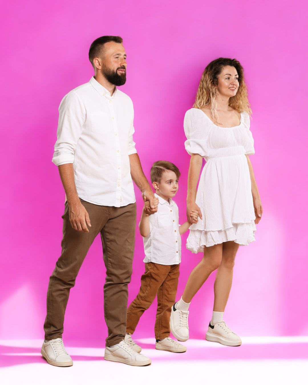 Una familia conformada por un hombre, una mujer y un niño, todos vestidos de blanco y sosteniéndose de las manos, en un fondo rosa.
