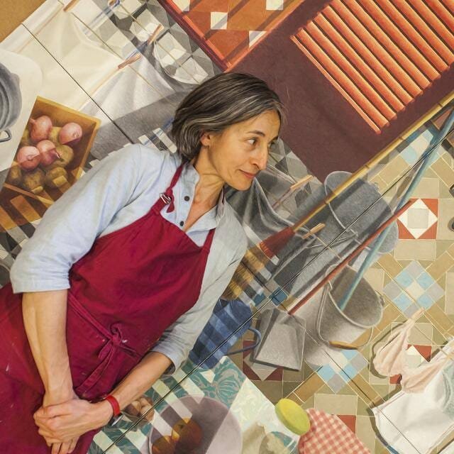 Eva - Cap de taller per a Made My Dia amb un davantal vermell sobre una camisa blau clar, dreta en una cuina amb un terra de rajoles de colors. Mira a la seva dreta amb una expressió pensativa. Hi ha productes de neteja i una cistella de cebes roses a prop.