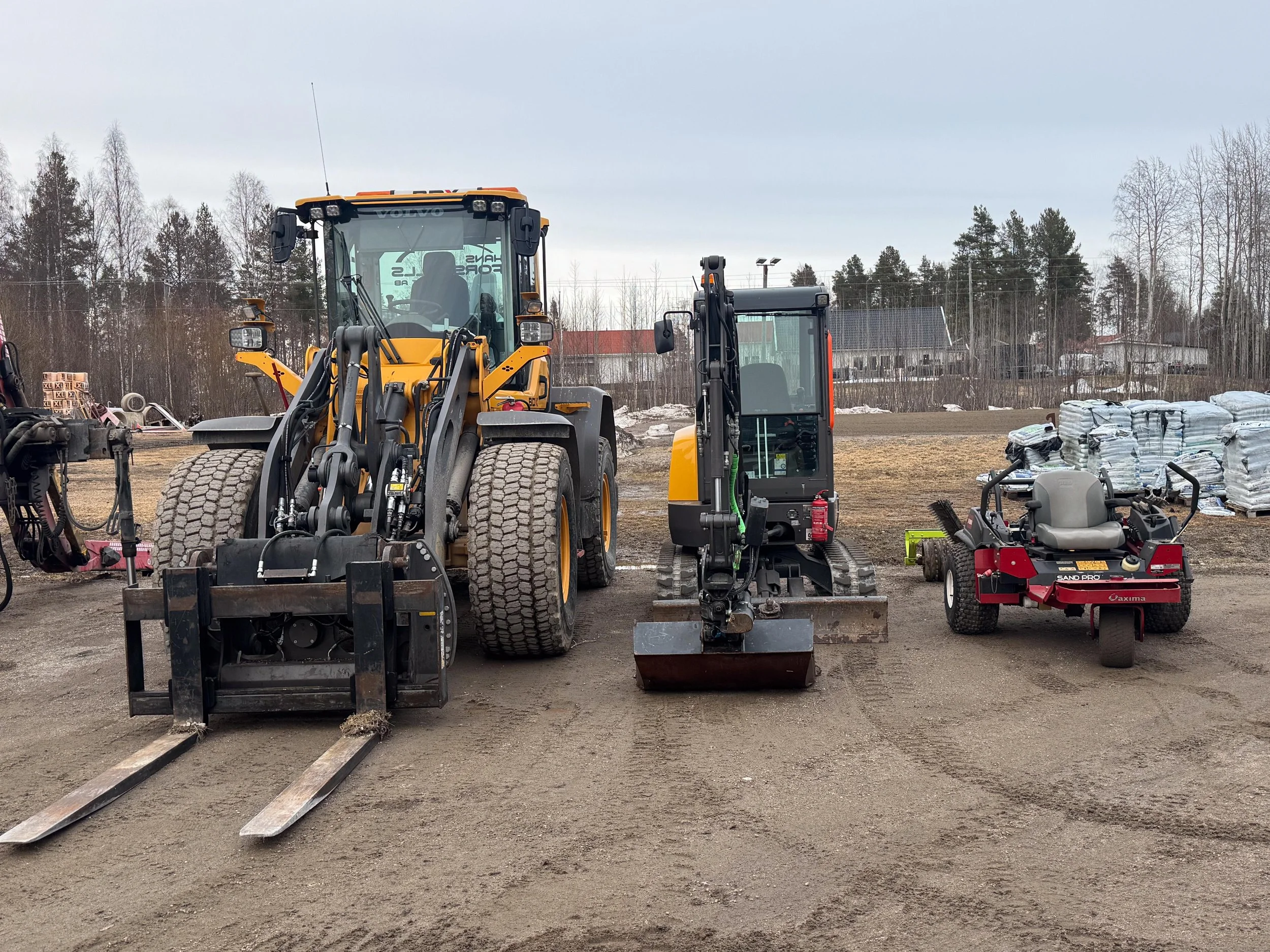 Tre olika typer av bygg- och markarbetesmaskiner på en byggarbetsplats, med träd och hus i bakgrunden.
