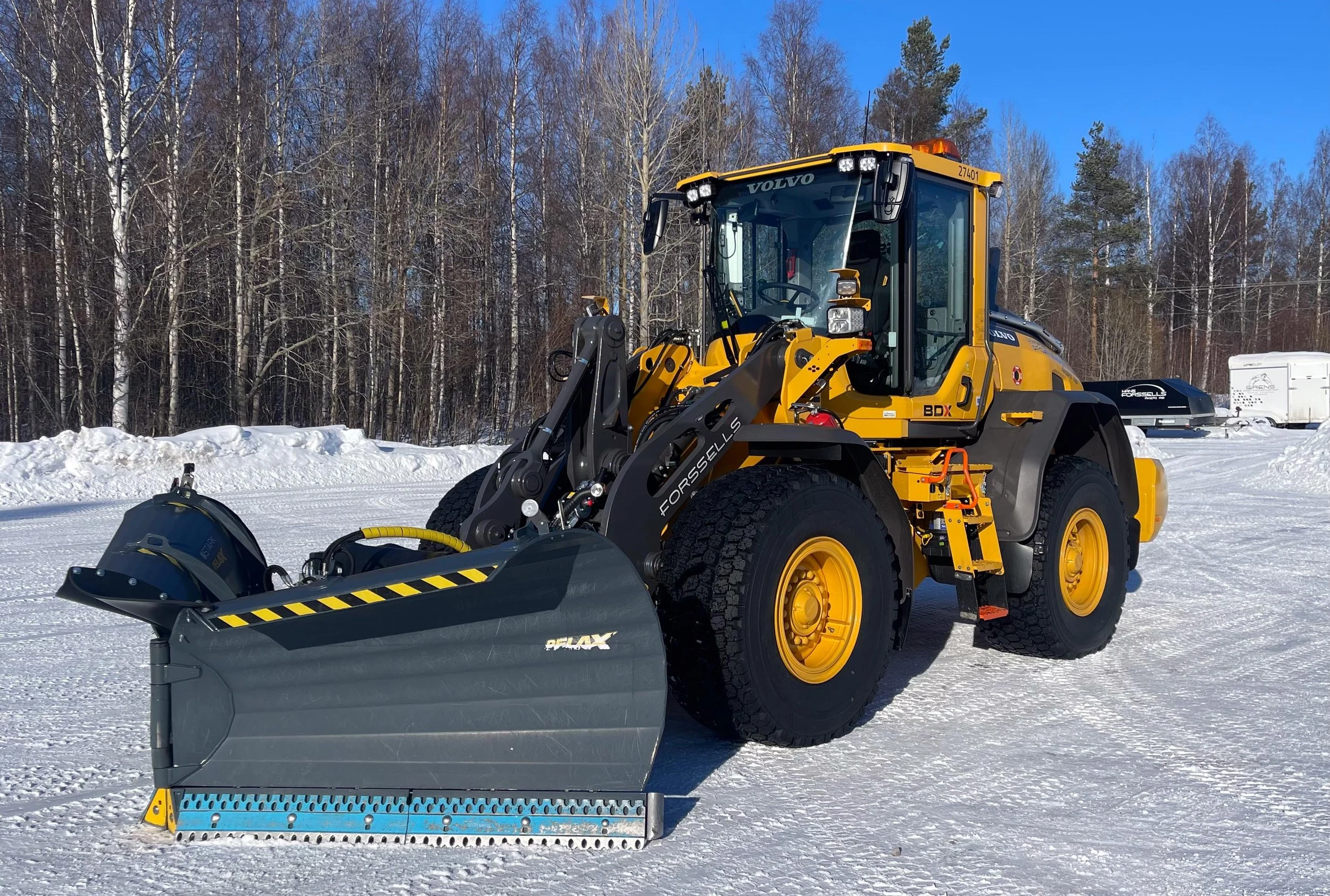 En gul och svart volvo snölastare på snöigt fält med träd i bakgrunden.