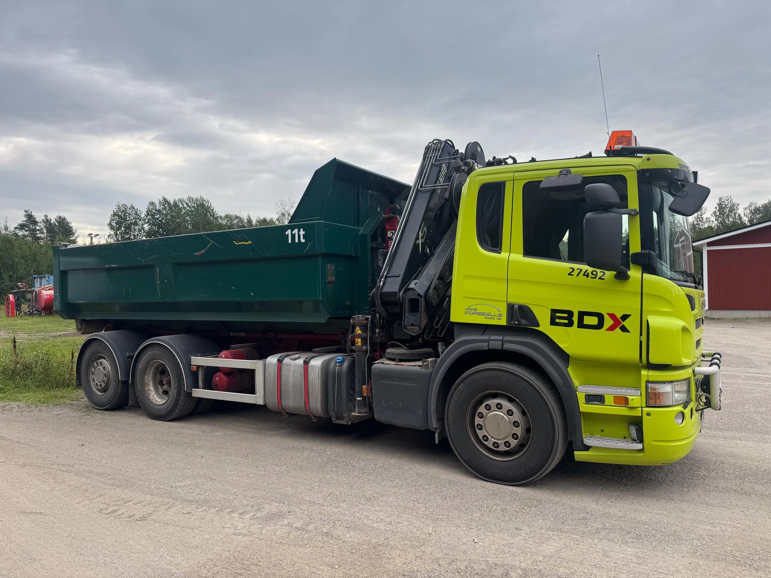En grön och gul lastbil med kranparaply, parkerad på en grusväg, mot en bakgrund av träd och en röd byggnad.
