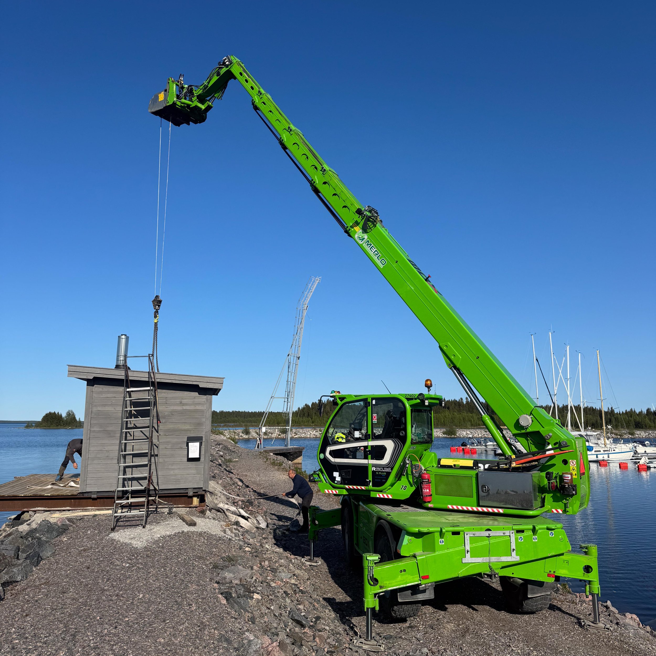En grön mobilkran lyfter en liten stuga eller chock ett landningsutrymme vid vattnet på en klar dag, med några personer som arbetar i närheten och båtar i bakgrunden.