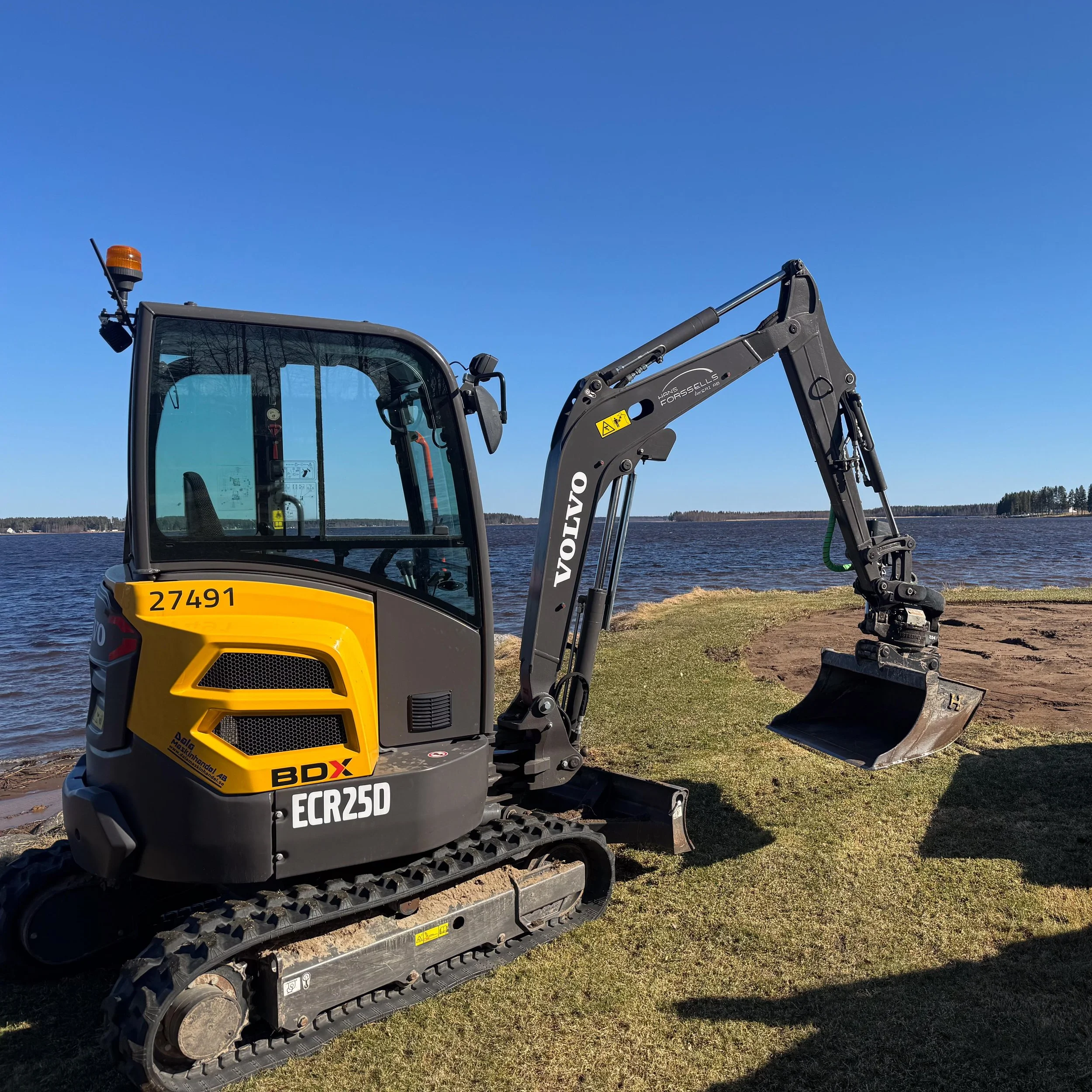 En liten grävmaskin, modell ECR25D, parkerad vid en sjö på en gräsyta.