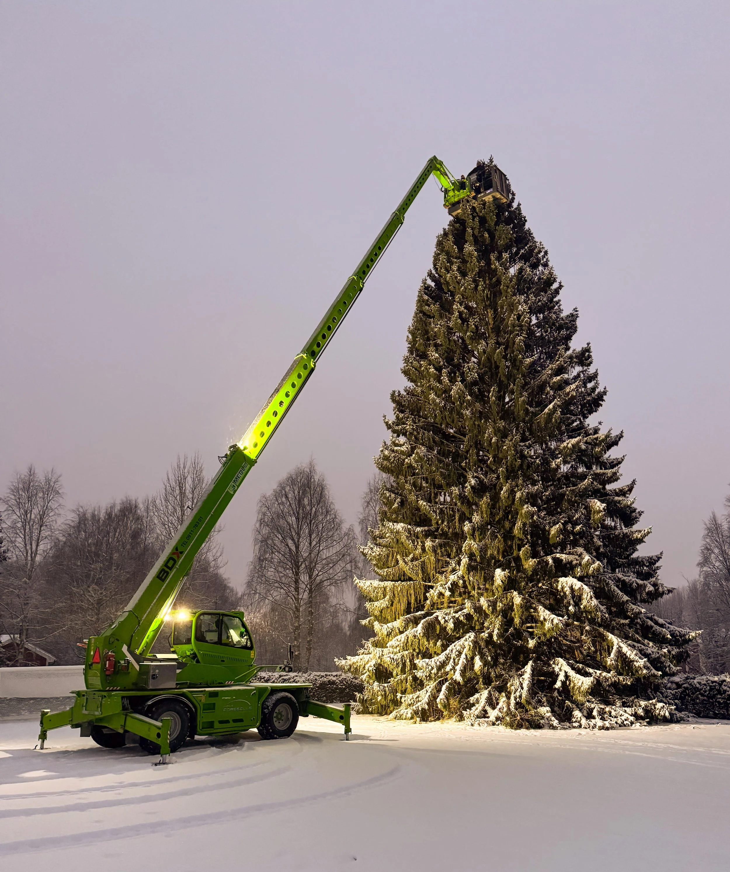 En grön mobilkran som lyfter en julgran i snöigt vinterlandskap.