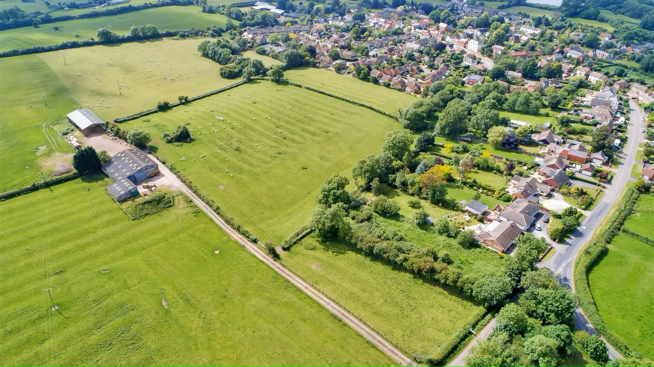 Aerial view of a residential neighborhood with green fields, trees, and houses, showing rural and suburban areas with open grassy spaces and farm buildings.
