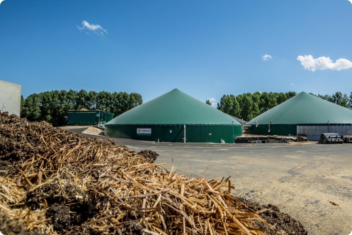 An anaerobic digestor (AD plant) outdoors under a blue sky with few clouds, and a pile of dried plant debris in the foreground.