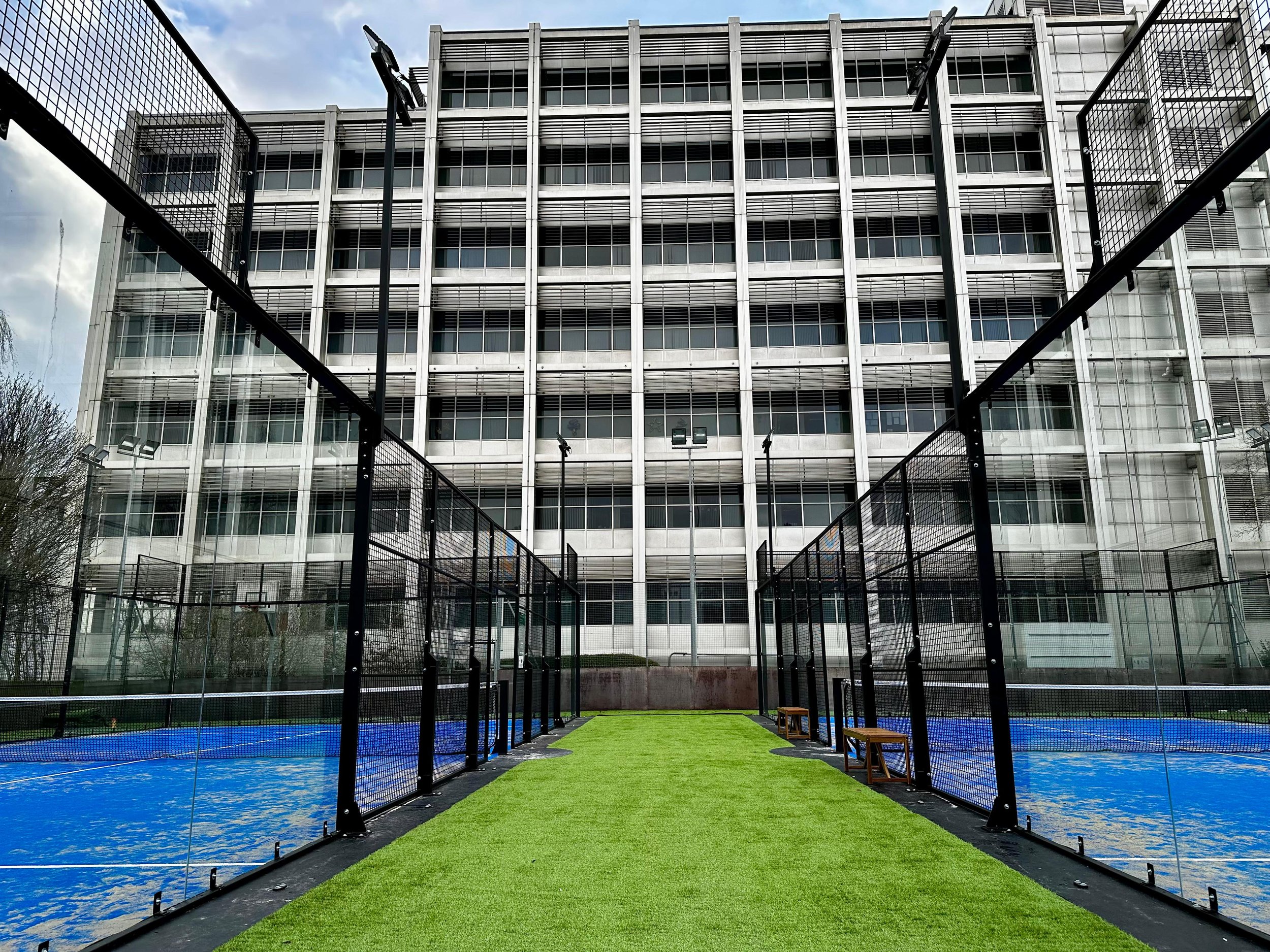 A multi-story modern residential or office building behind a padel tennis court with blue surfaces and a green artificial turf pathway. Benches are on either side of the pathway.