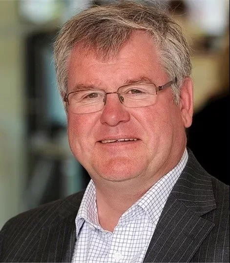 A middle-aged man with glasses, gray hair, wearing a dark suit and a light checkered shirt, smiling at the camera.
