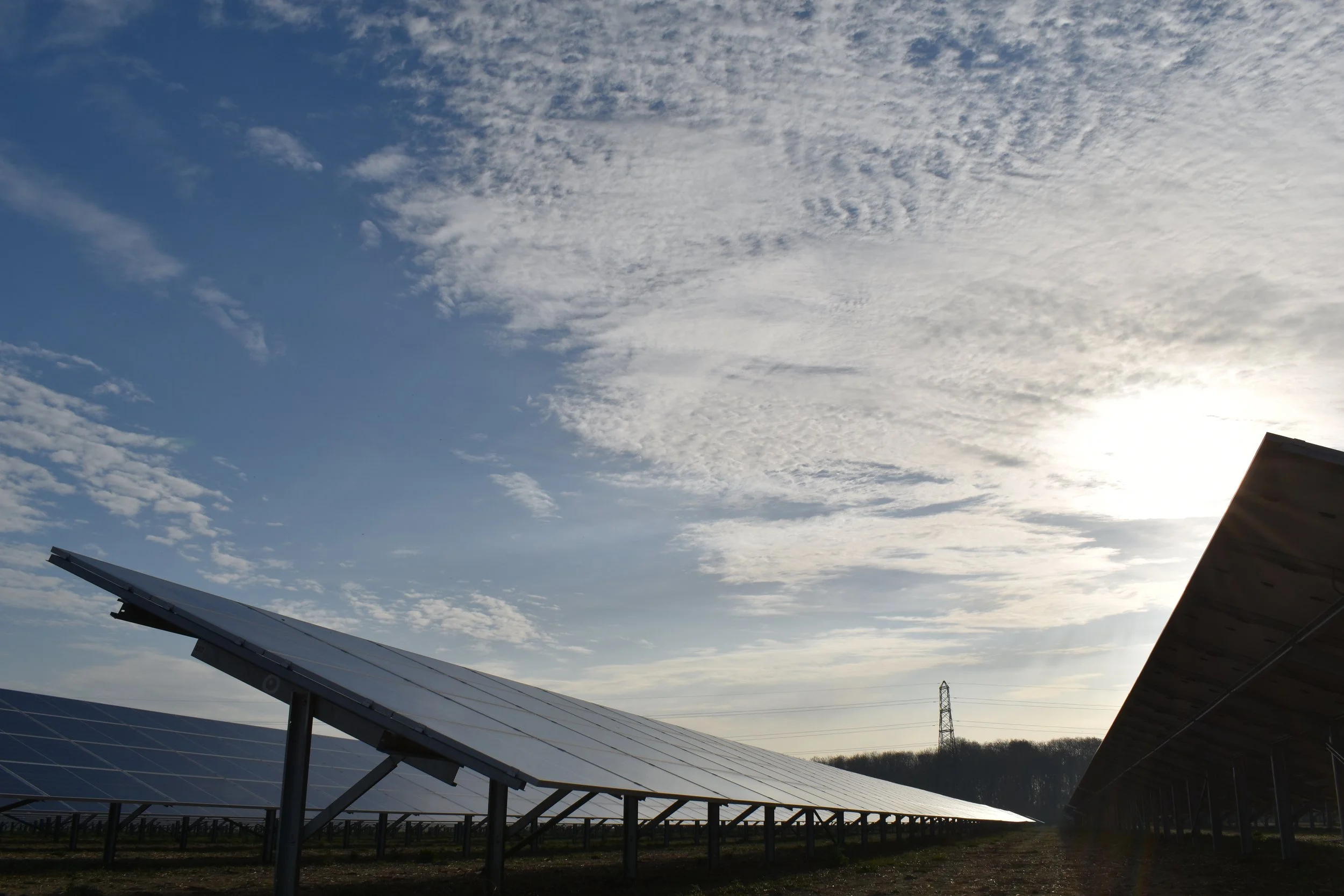 Solar panels in a field with a partly cloudy sky and the sun shining from behind them.
