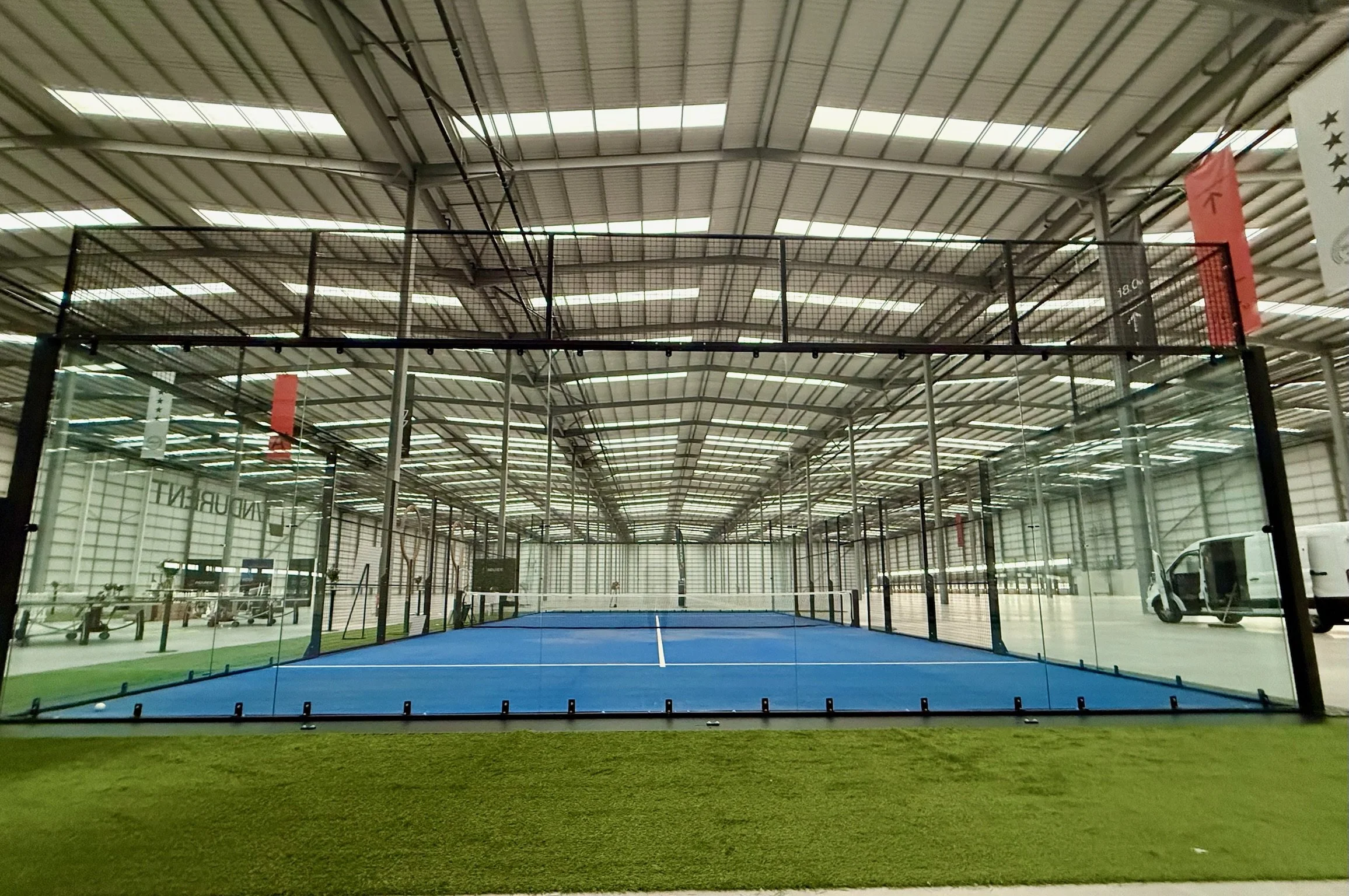 Indoor sports facility with a blue tennis or padel court enclosed by glass walls and fencing, with a concrete floor surrounding it.