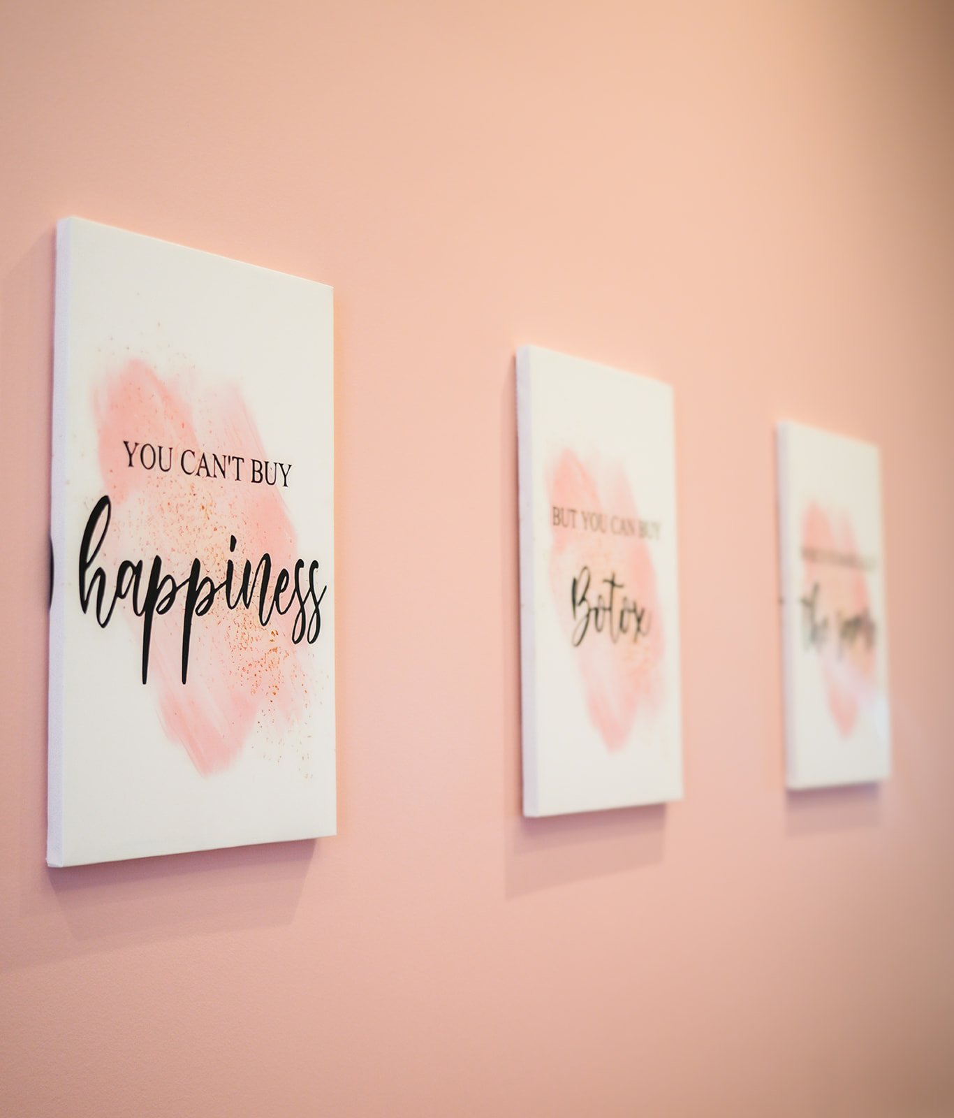 Three framed art prints hanging on a pink wall. The prints have pastel watercolor backgrounds with motivational quotes about happiness and self-care.