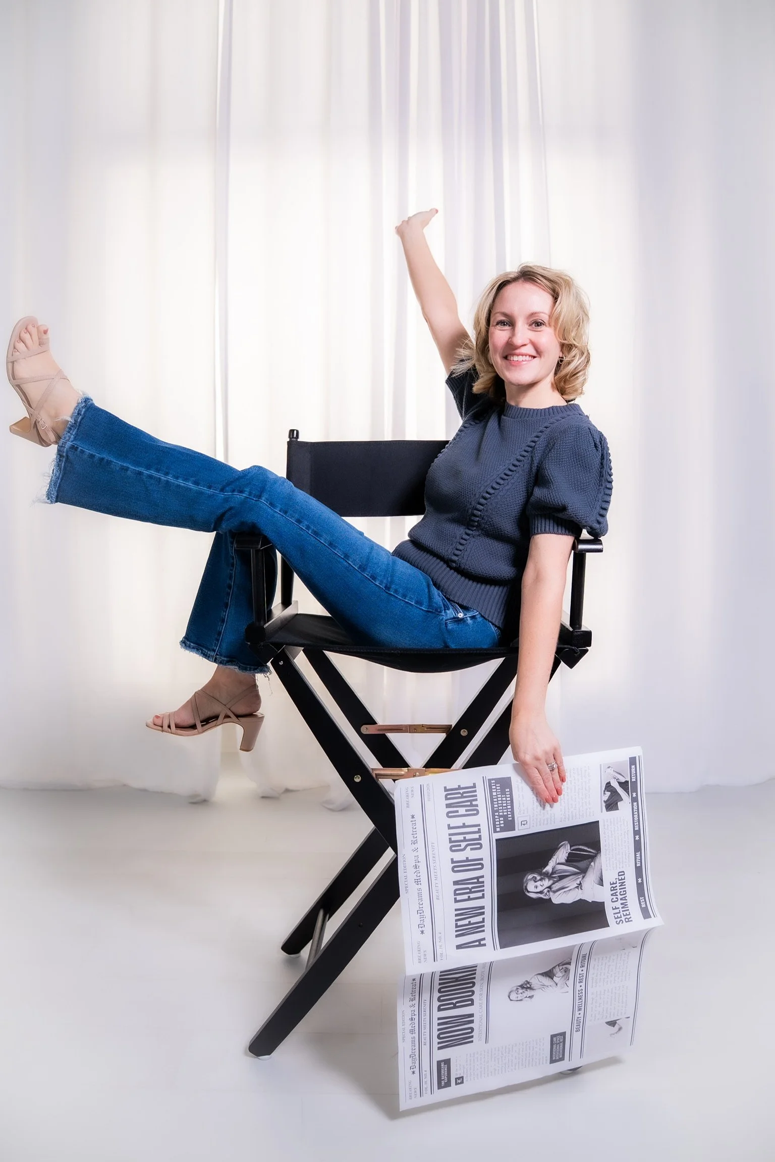 A woman with blond hair sitting on a black chair, smiling, wearing a dark blue sweater and blue jeans, with one leg resting on the chair and the other touching the floor. She is holding a newspaper on the floor with her right hand, and her left arm is raised above her head. The background is white with sheer curtains, and the floor is white.