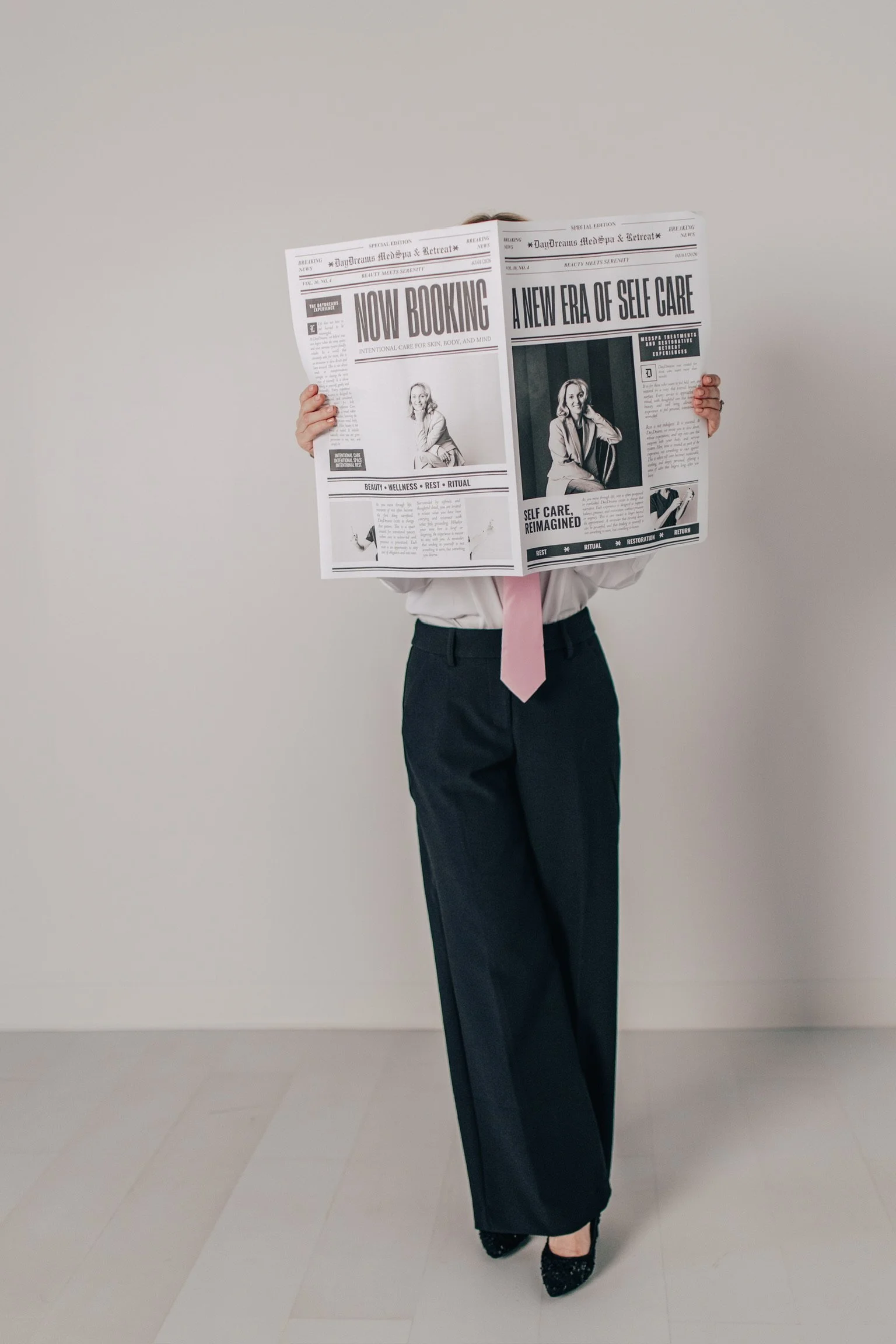 Person dressed in a white shirt, pink tie, dark pants, and black shoes holding a newspaper in front of their face, standing against a plain light-colored wall.